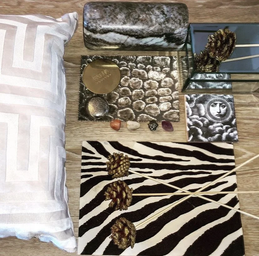 Decorative display on a wooden surface featuring a pillow, marble and metallic objects, a glass with a snack, a mask, a framed face, a zebra striped tray with pinecones, and several small decorative items and stones.