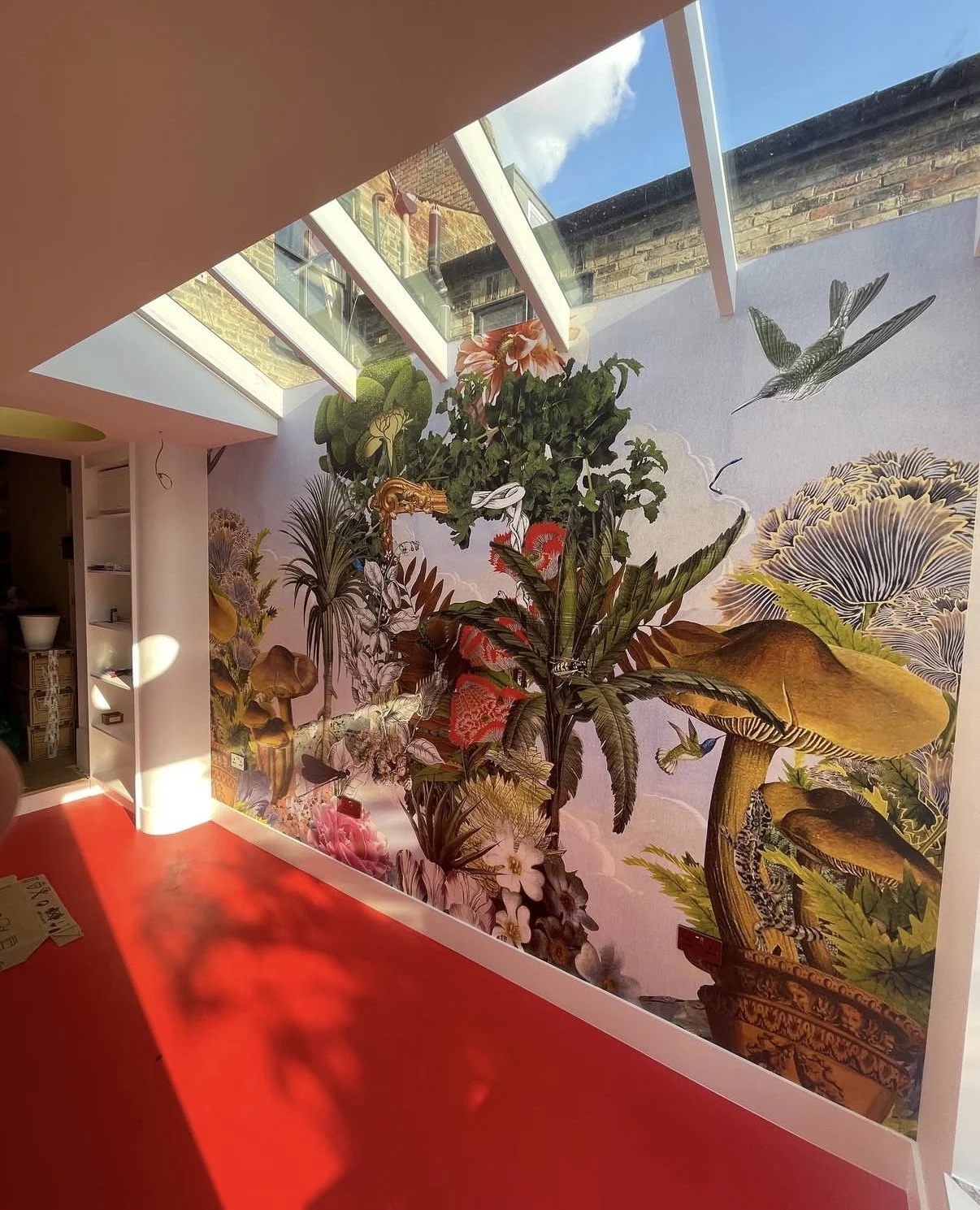 Interior space with large wall mural of mushrooms, plants, and birds, red carpet flooring, and skylights letting in natural light.