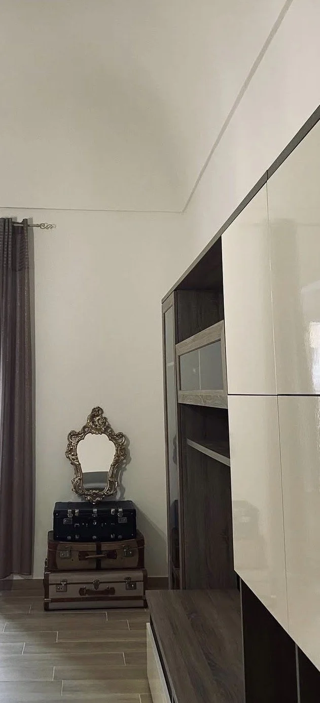 Corner of a room showing a vintage mirror, a small stack of suitcases, a dark wood cupboard with glass doors, a beige wall, and a window with dark curtains.