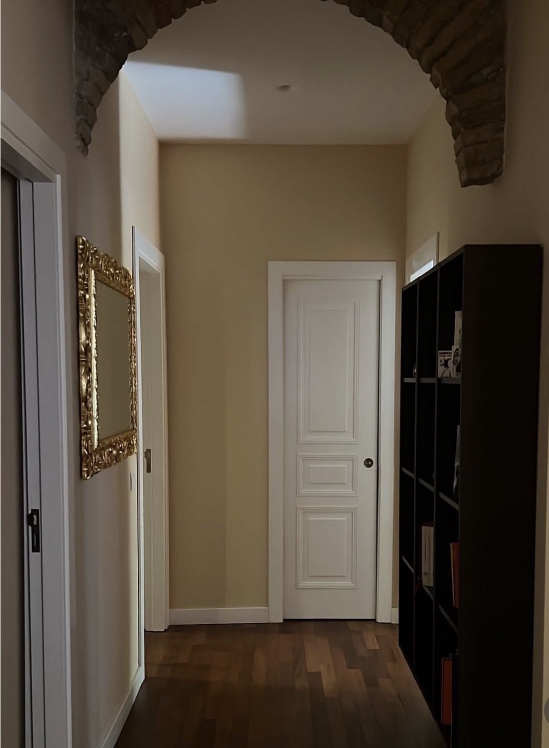 A hallway with beige walls, a hardwood floor, a white door with decorative trim, a black bookshelf on the right, and a gold-framed mirror on the left wall. An exposed brick arch is visible at the top of the hallway.