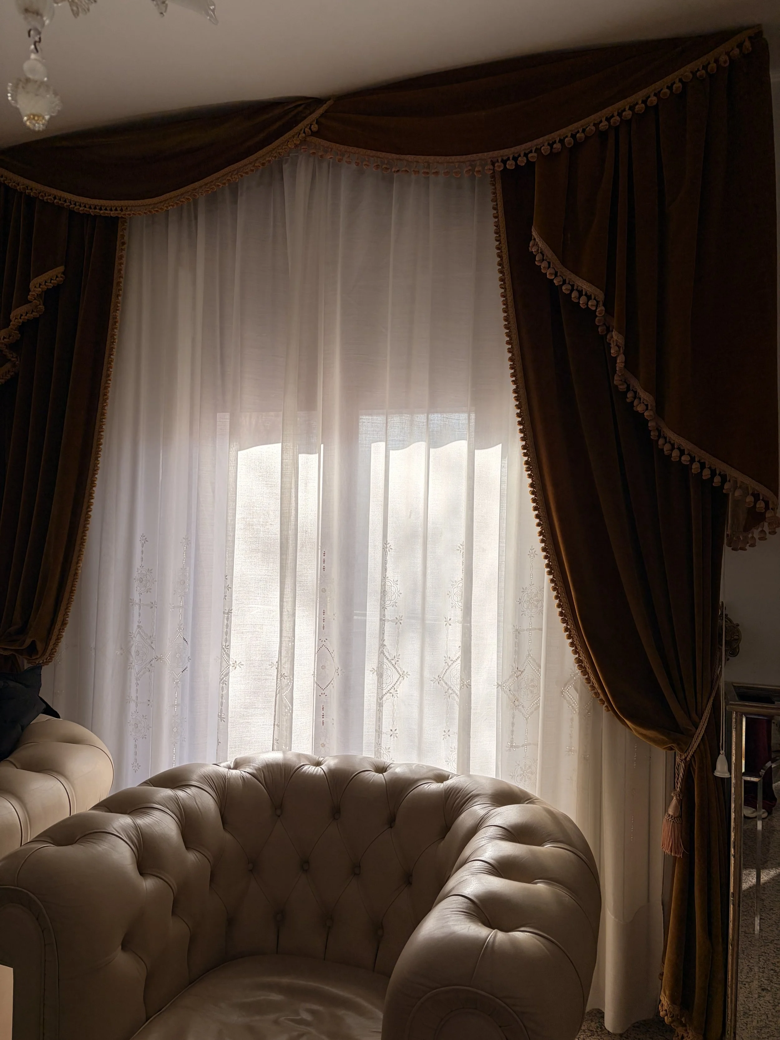 Living room window with sheer white curtains and dark brown drapes with tassel trim, and beige tufted leather armchair.