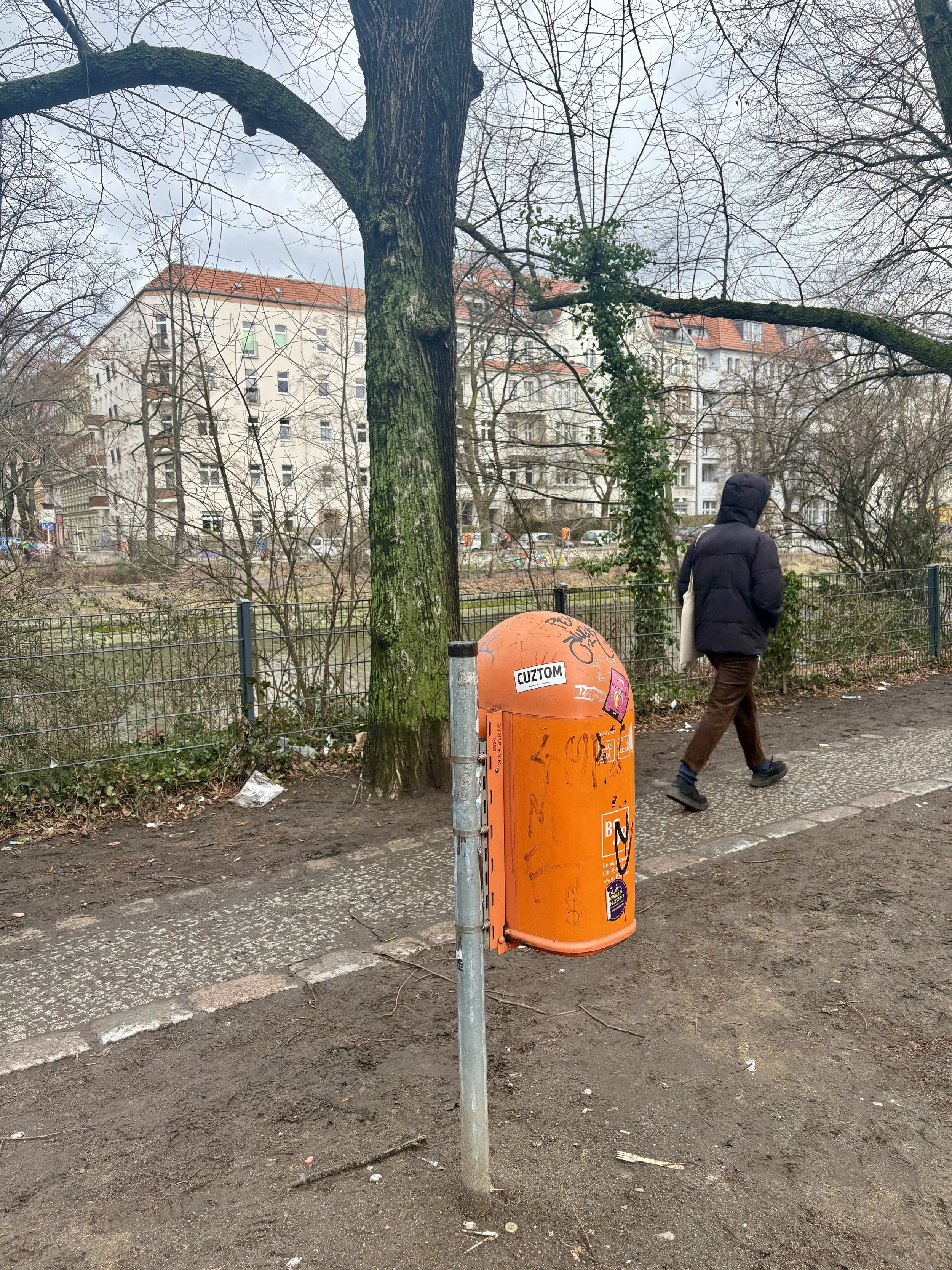 An orange mailbox stands on a sidewalk with a person in a black coat walking past it, behind a tree in a park, with apartment buildings in the background.