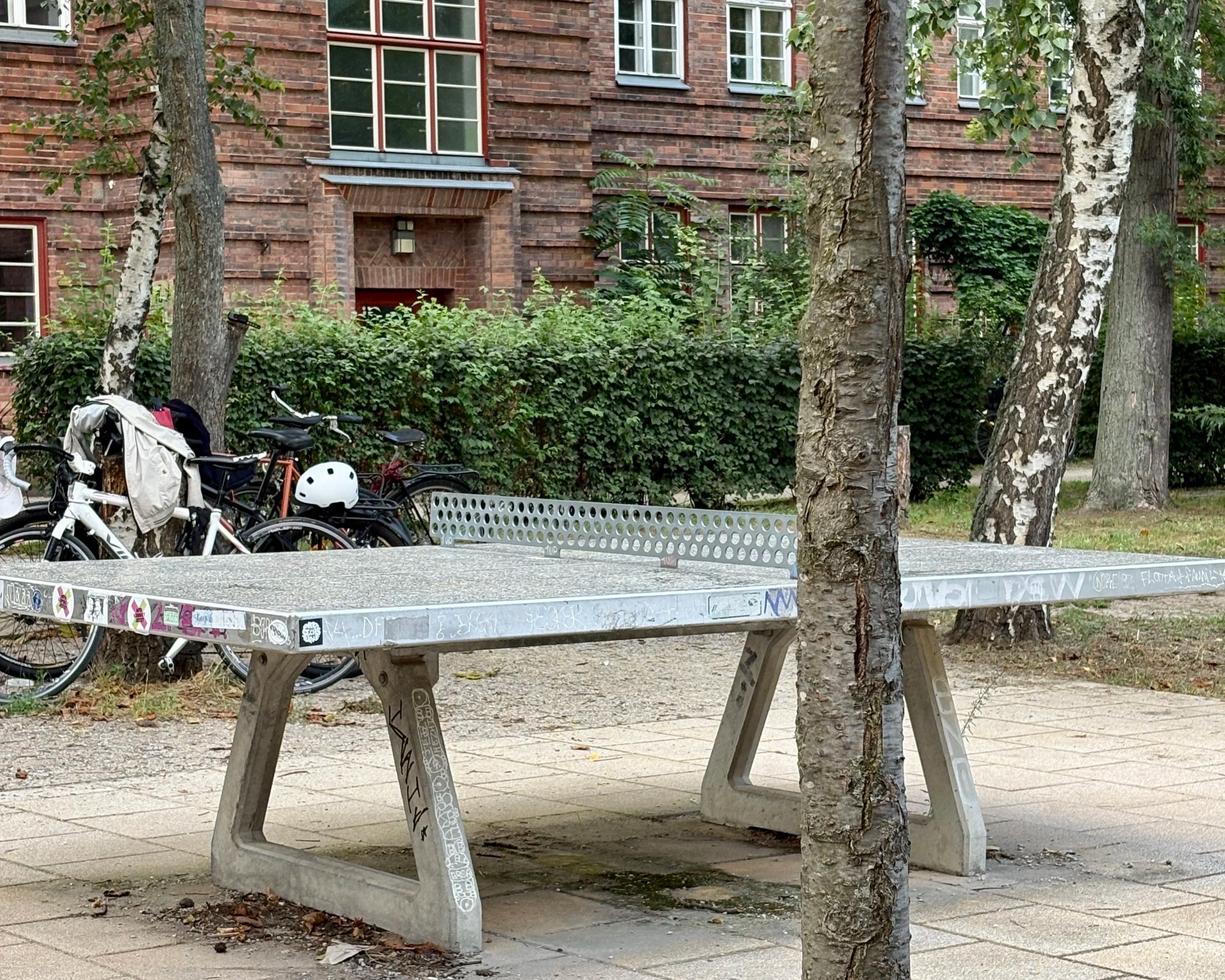 An outdoor table tennis table with graffiti on its side, located on a sidewalk in a park with trees, benches, bicycles, and a brick building in the background.