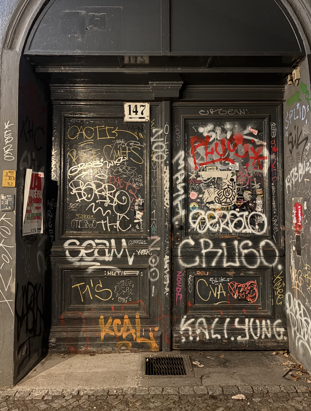 A black metal door covered in colorful graffiti, stickers, and tags with the house number 147 above the door, set in a stone archway.