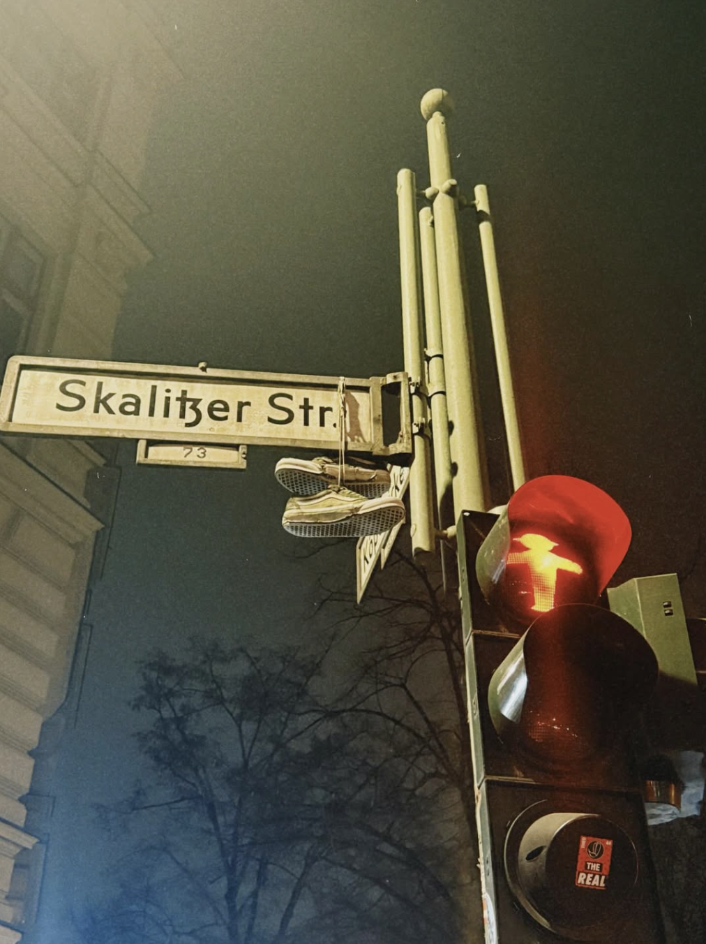 Street sign reading 'Skalitzer Str.' with a pedestrian crossing signal showing a red hand. Shoes are hanging from the street sign post. Trees and building facades are visible in the background on a foggy night.