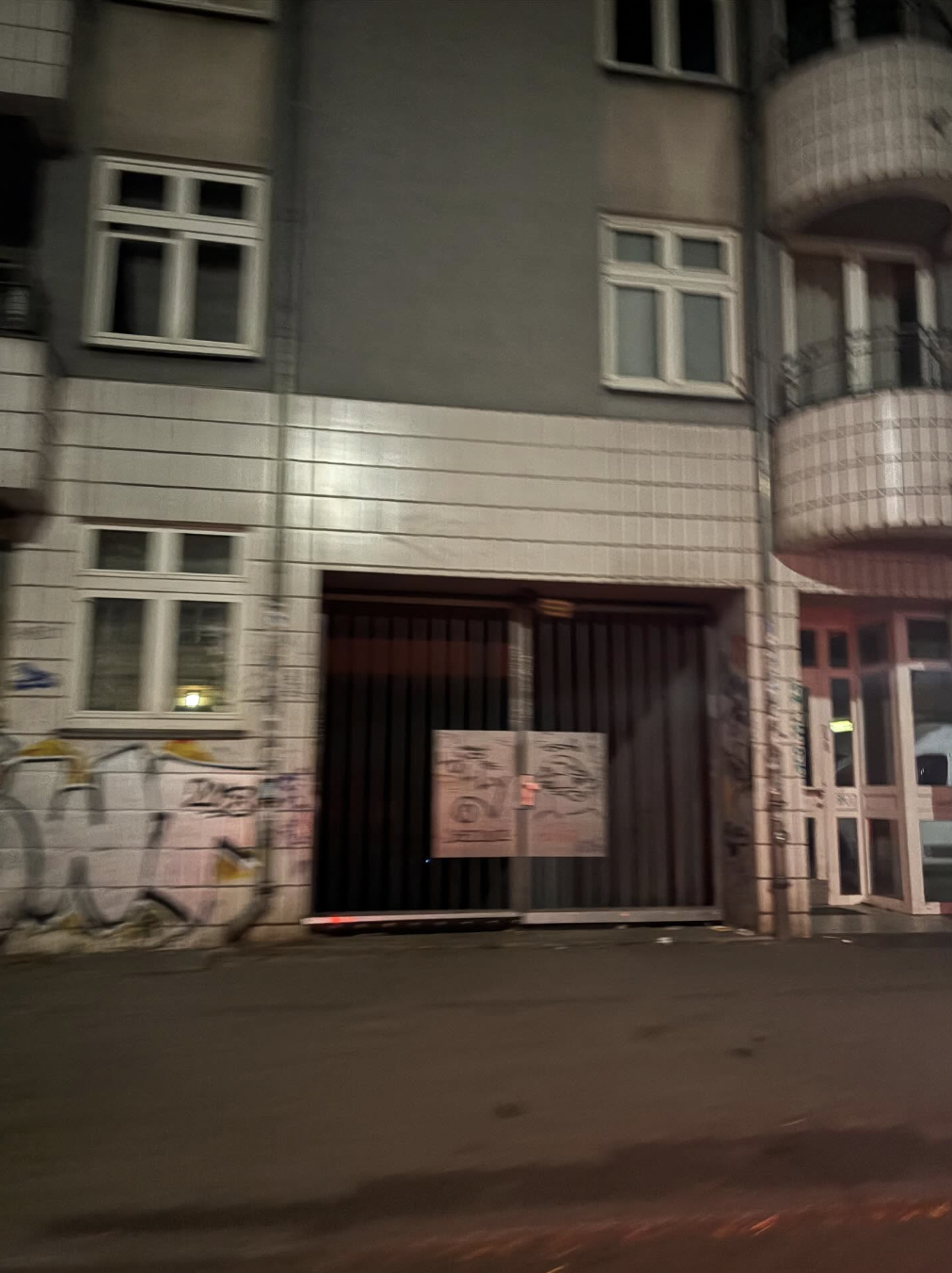 Blurred nighttime photo of an apartment building with graffiti on the wall, several windows, and a large black gate at the entrance.