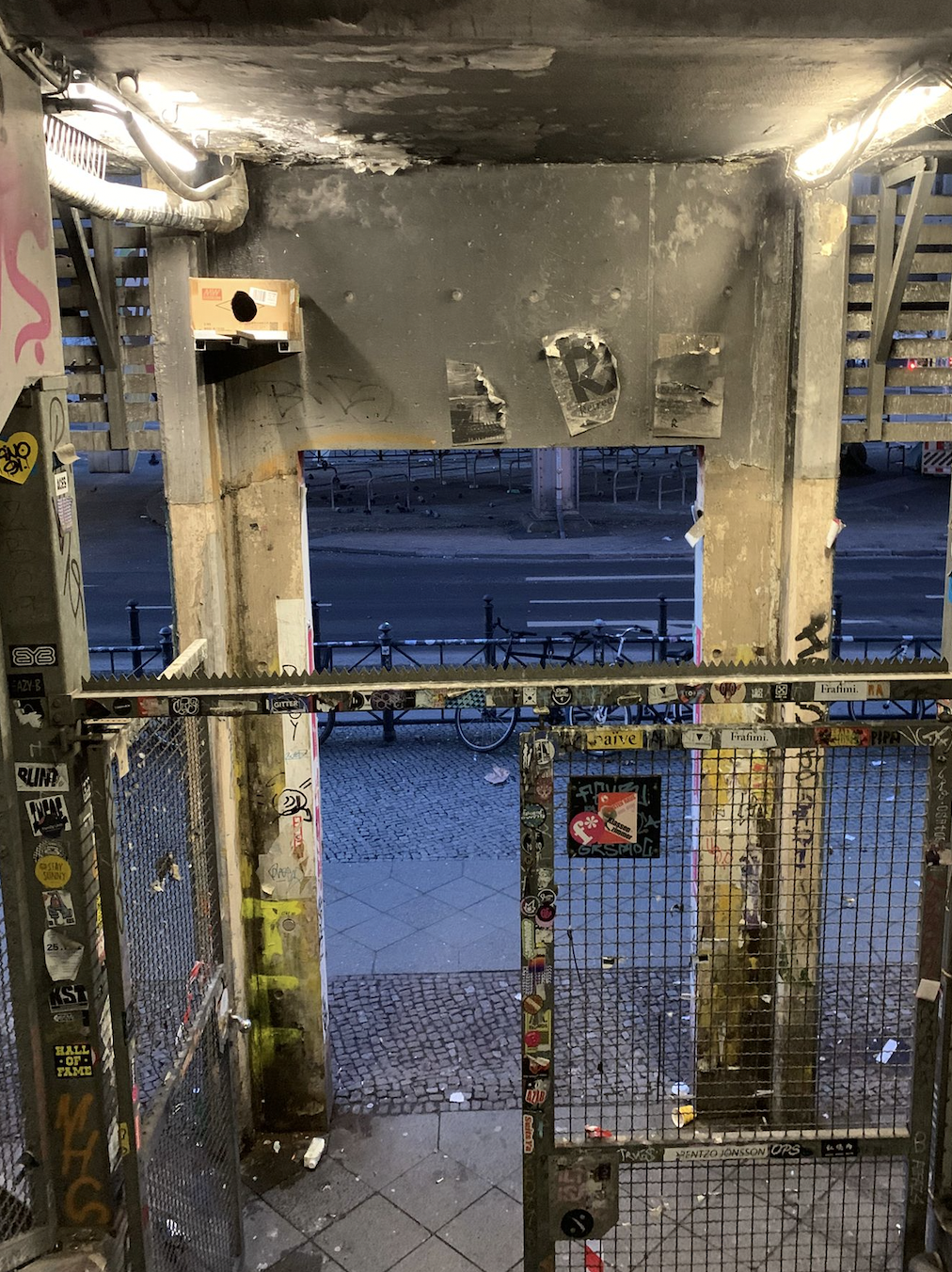 View from inside a graffiti-covered underpass or tunnel with a metal fence at the entrance, looking out onto a street with a sidewalk and bicycle rack, illuminated by overhead lights.