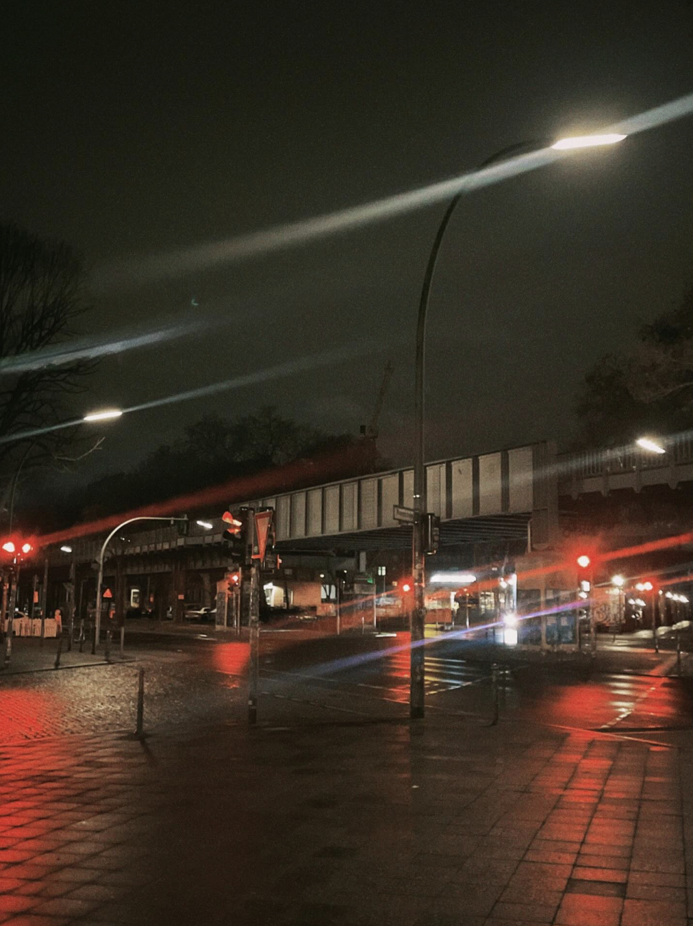 Nighttime city street scene with wet pavement reflecting red traffic lights, street lamps illuminating the area, and an elevated train track crossing above. There are trees and some buildings in the background.