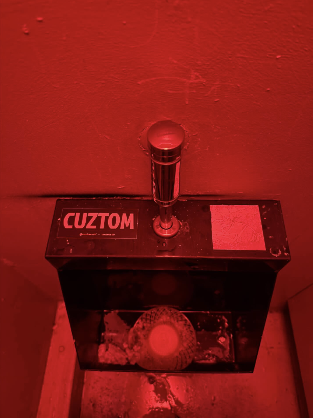 A black box with a sticker that reads 'CUZTOM,' a red sticker with social media handles, and a metal spring-loaded foot pedal on top, all illuminated by red lighting.
