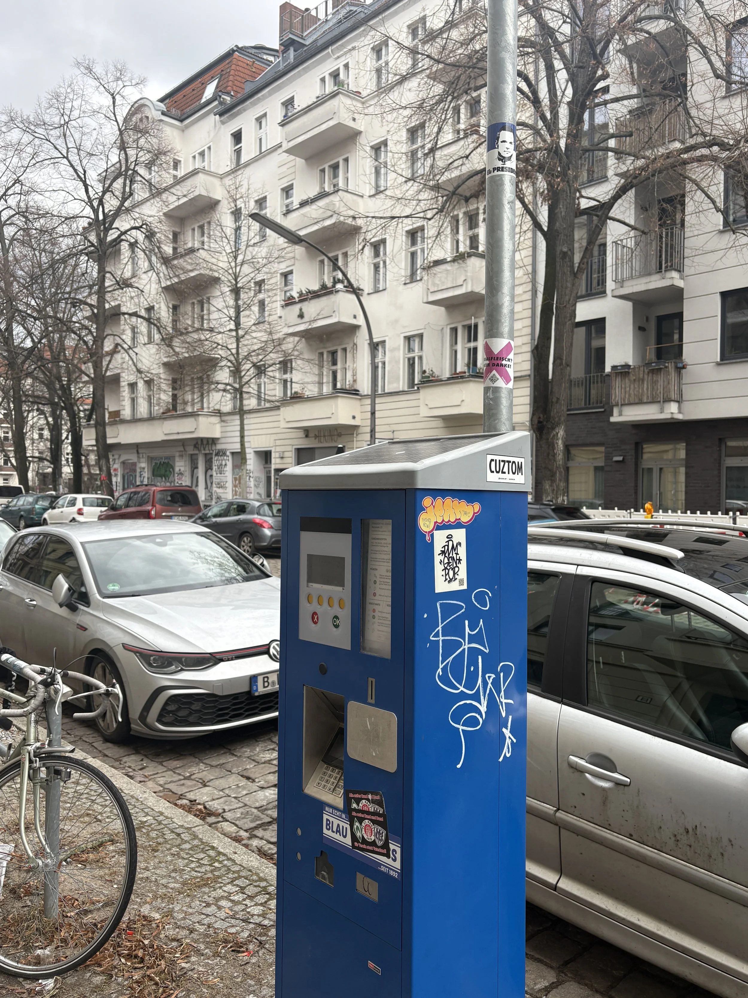 Blue parking meter on a city sidewalk with graffiti and stickers, parked cars, bicycles, and residential buildings with balconies in the background.