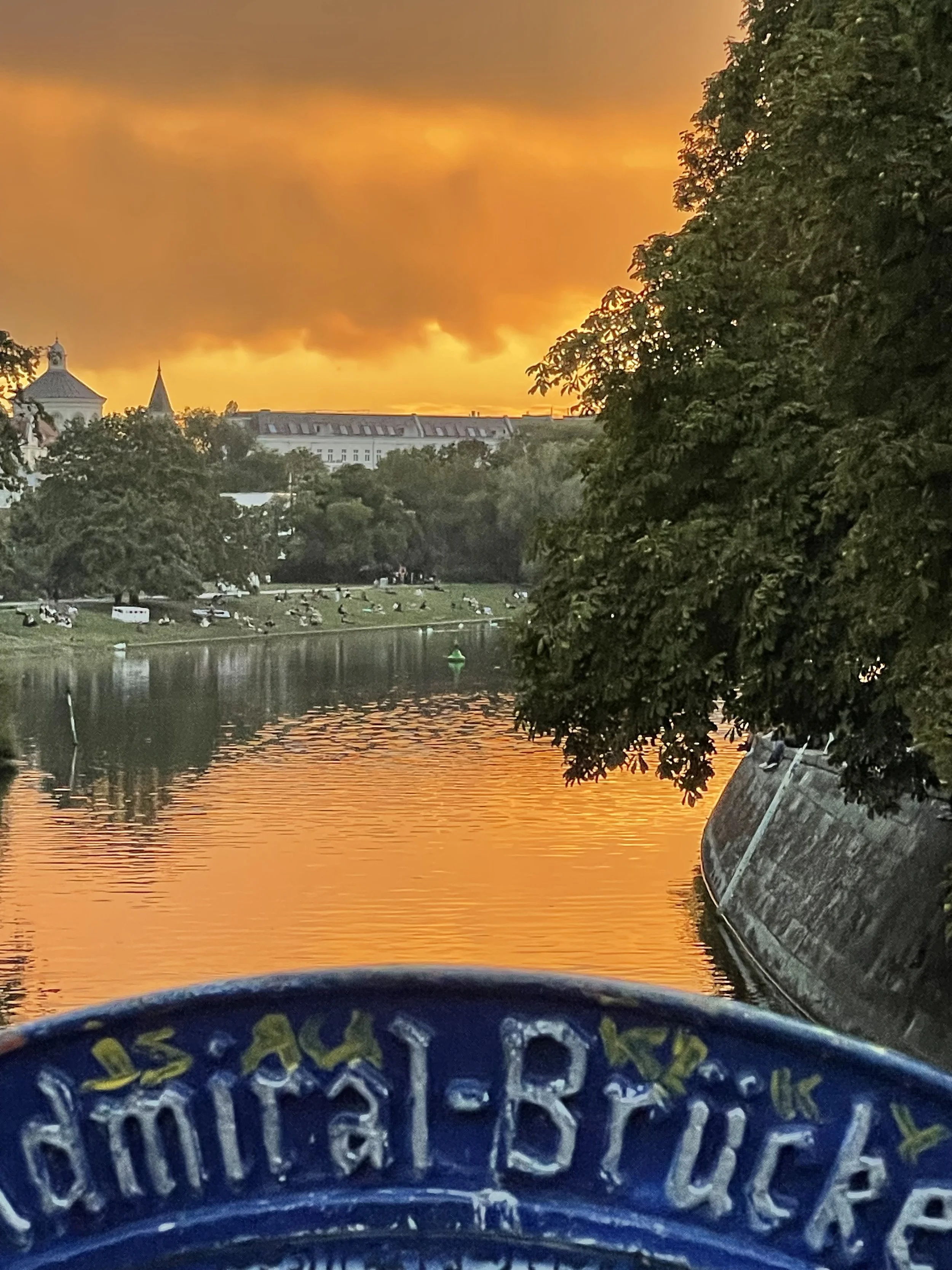 Sunset over a river with a brick building and trees on the bank, and a blue object in the foreground.