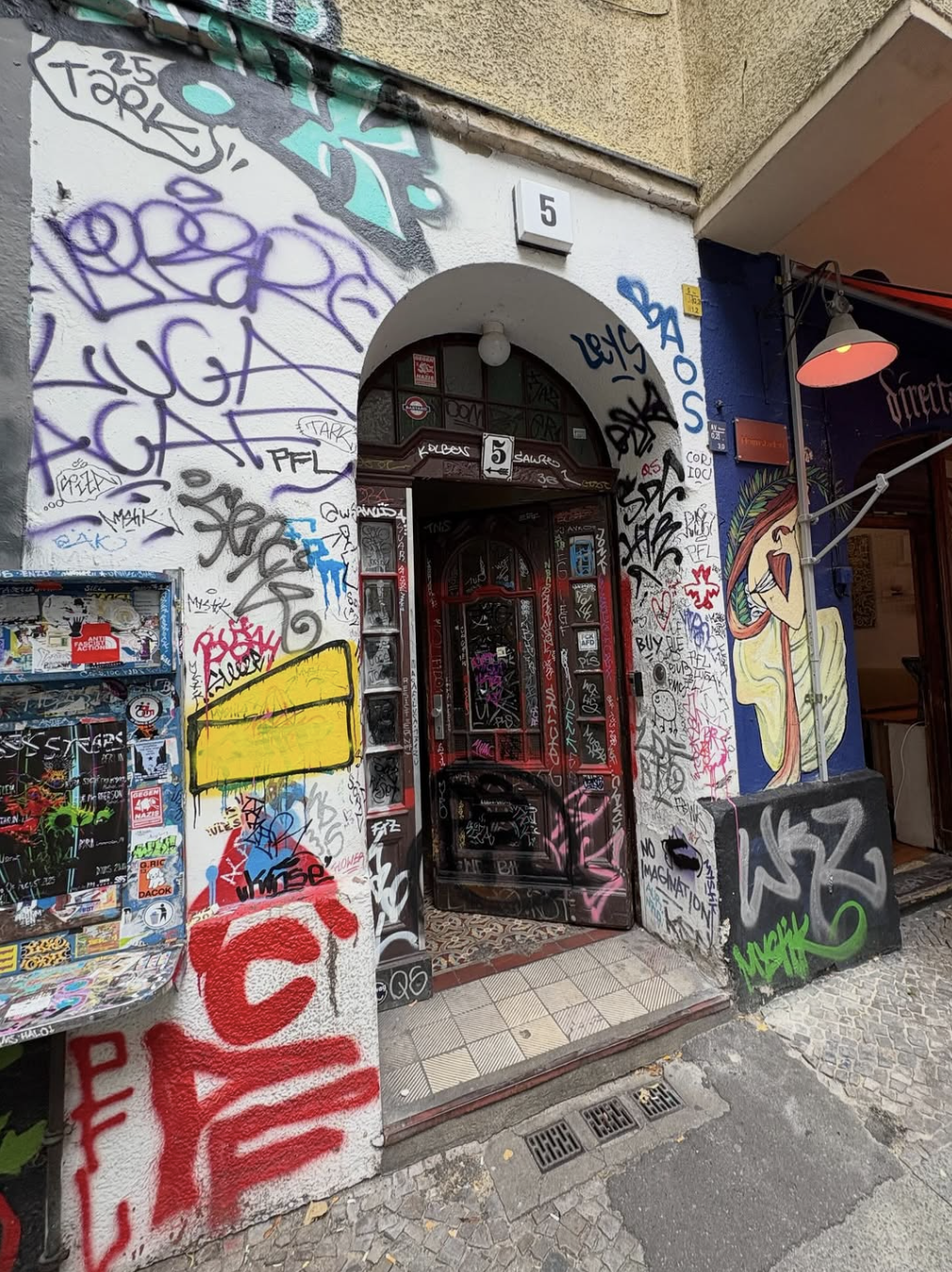 Graffiti-covered building entrance with a black door, small sign with the number 5, and colorful street art. The scene shows a blue wall and a mural of a woman on the right.