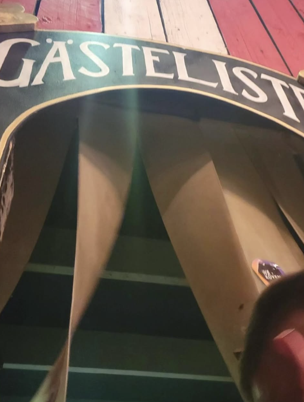 A green and black skateboard deck with a yellow border and the word "GASTELIST" visible on the top edge, placed on a wooden surface with painted red and tan planks.