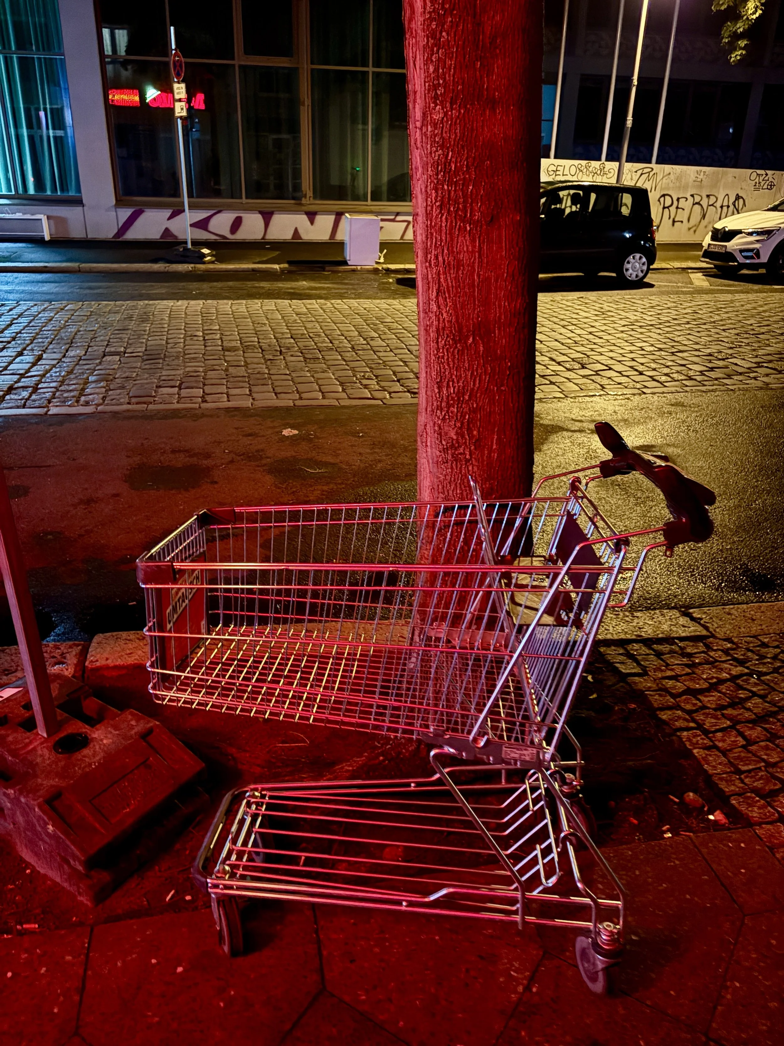 An empty shopping cart overturned on a sidewalk at night, illuminated by red and yellow streetlights, next to a tree and in front of a building with graffiti.