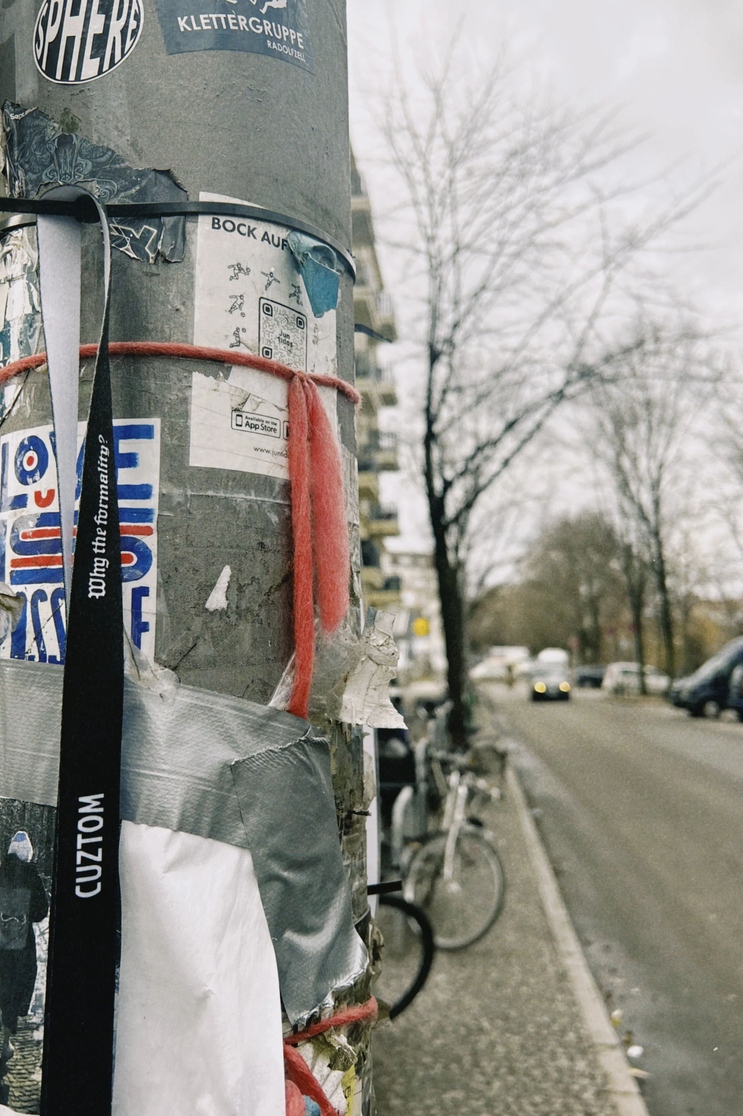 Close-up of a pole covered with stickers, posters, and a red string, with a street and parked bicycles in the background.