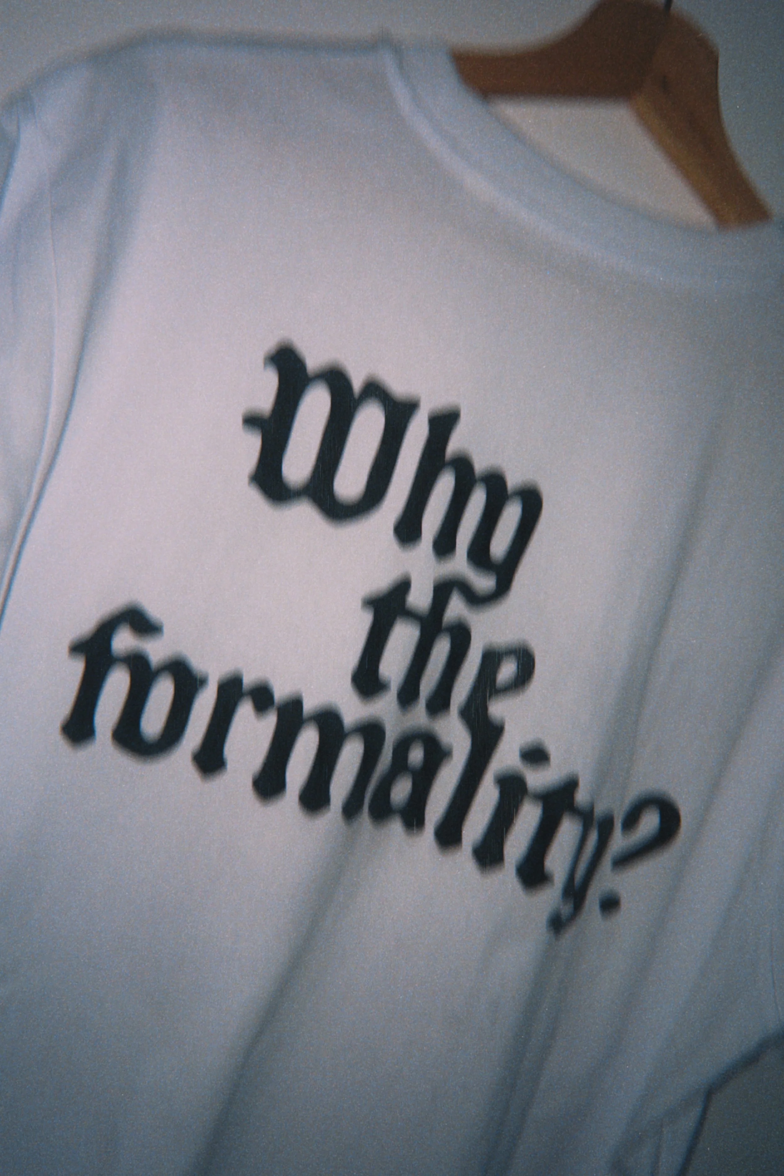 White t-shirt with the text 'why do I do this' in black, stylized font, hanging on a wooden hanger.