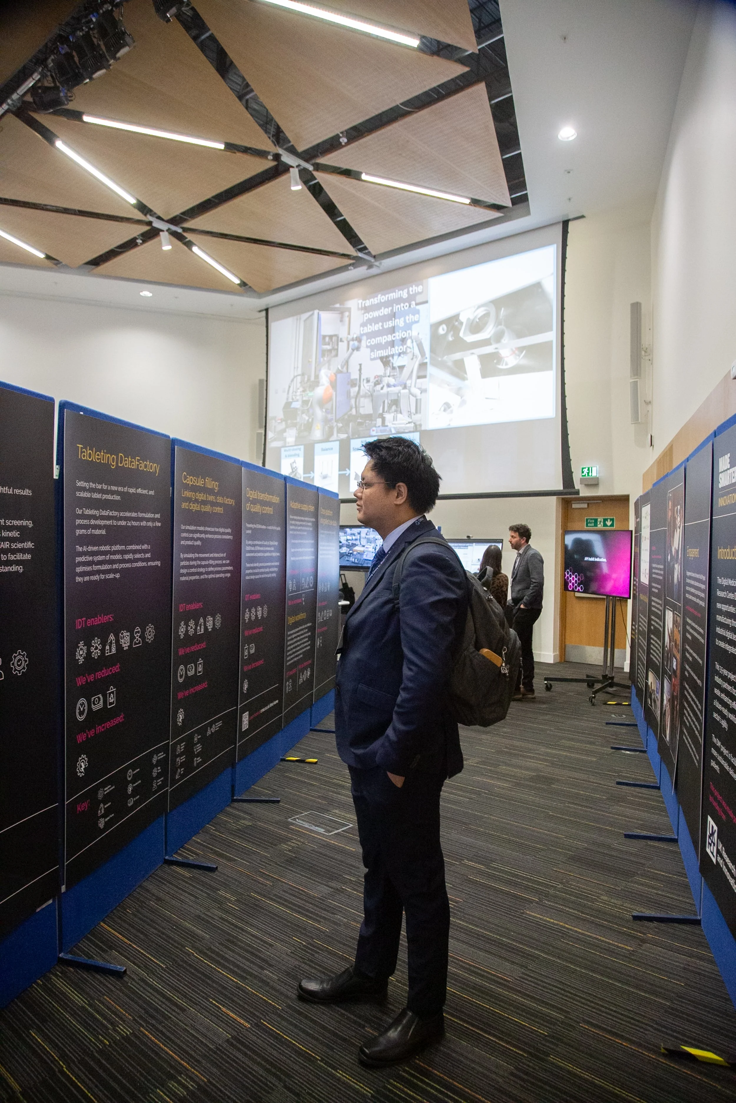 A man in business attire standing in front of informational display boards at a conference or exhibition.