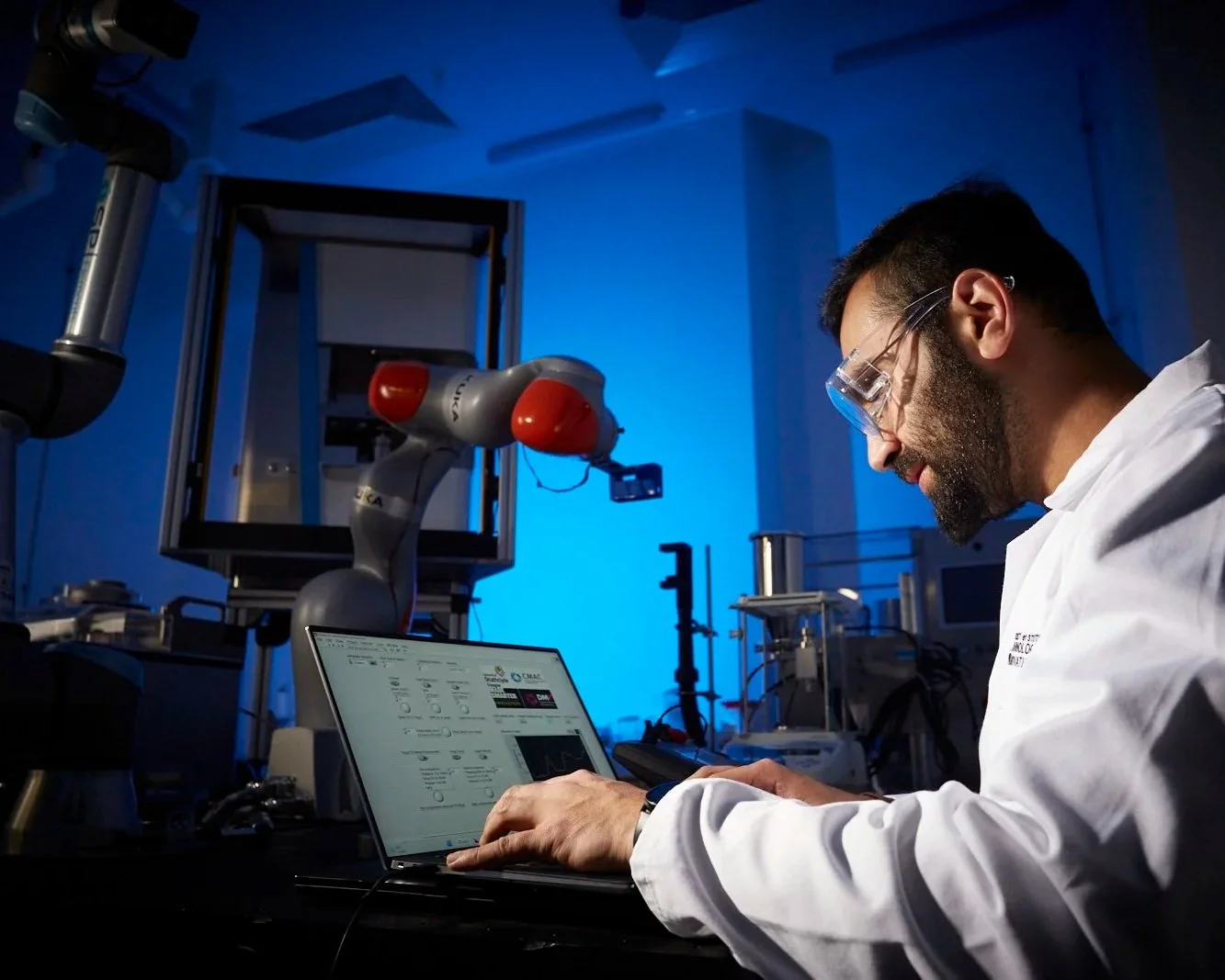 A scientist working in a lab, using a laptop, with a robotic arm in the background.