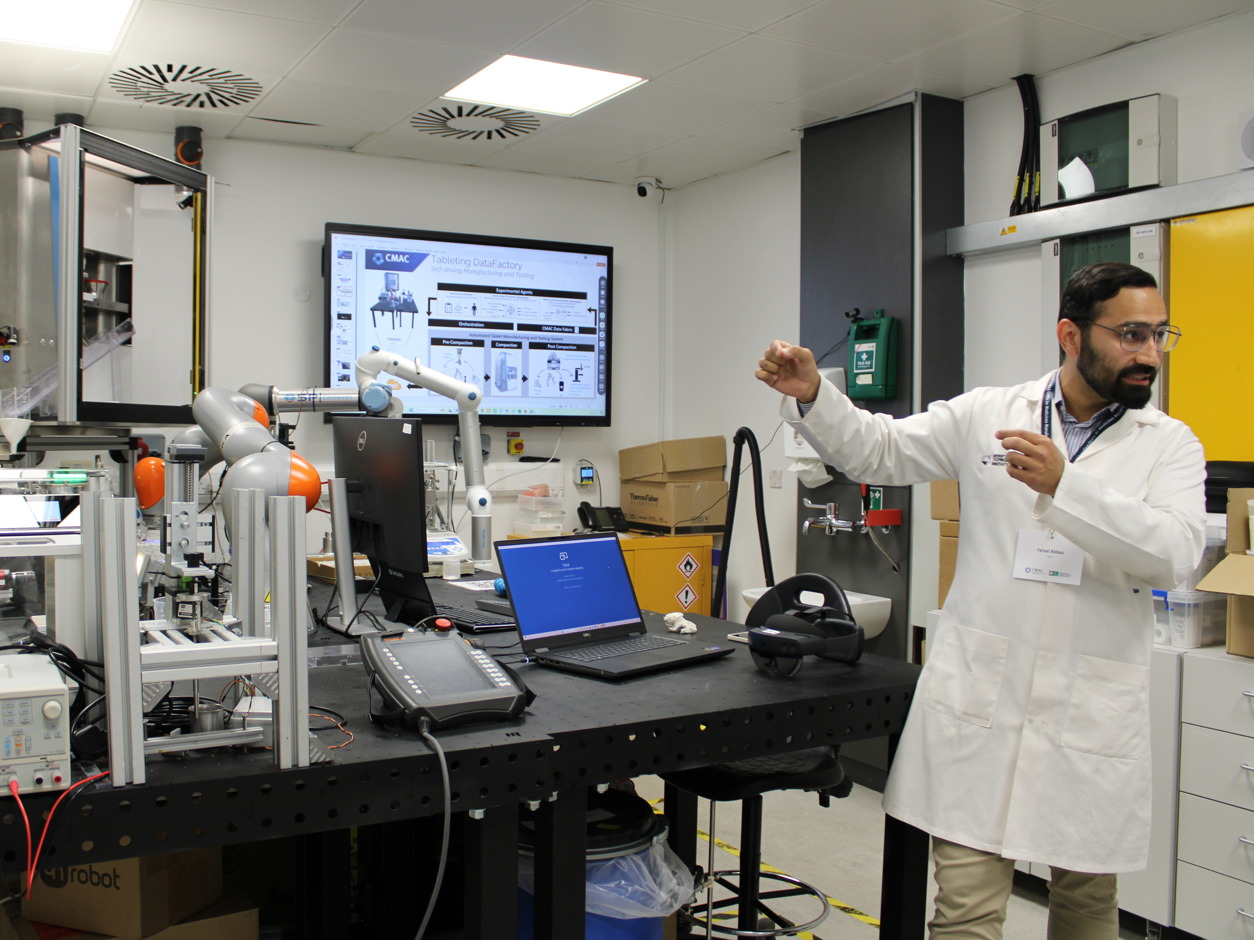 A man in a white lab coat explaining a robotic arm and a display screen showing a diagram related to data factory and testing in a laboratory setting with various equipment and tools.