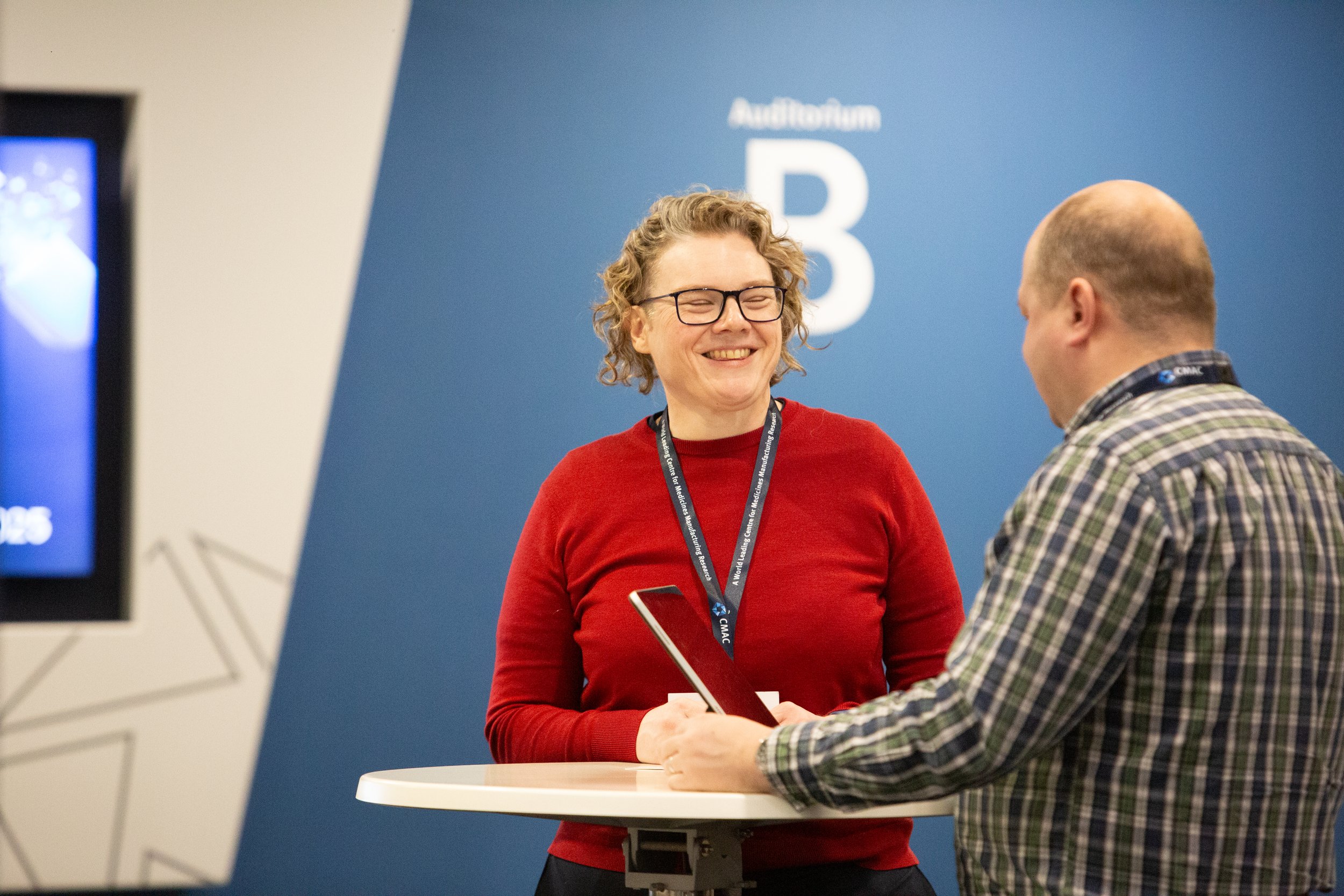 Two people, a woman with curly blonde hair and glasses wearing a red sweater and a man with a baseball cap and checkered shirt, are smiling and talking during a professional event or conference, standing around a small round table with conference badges visible.