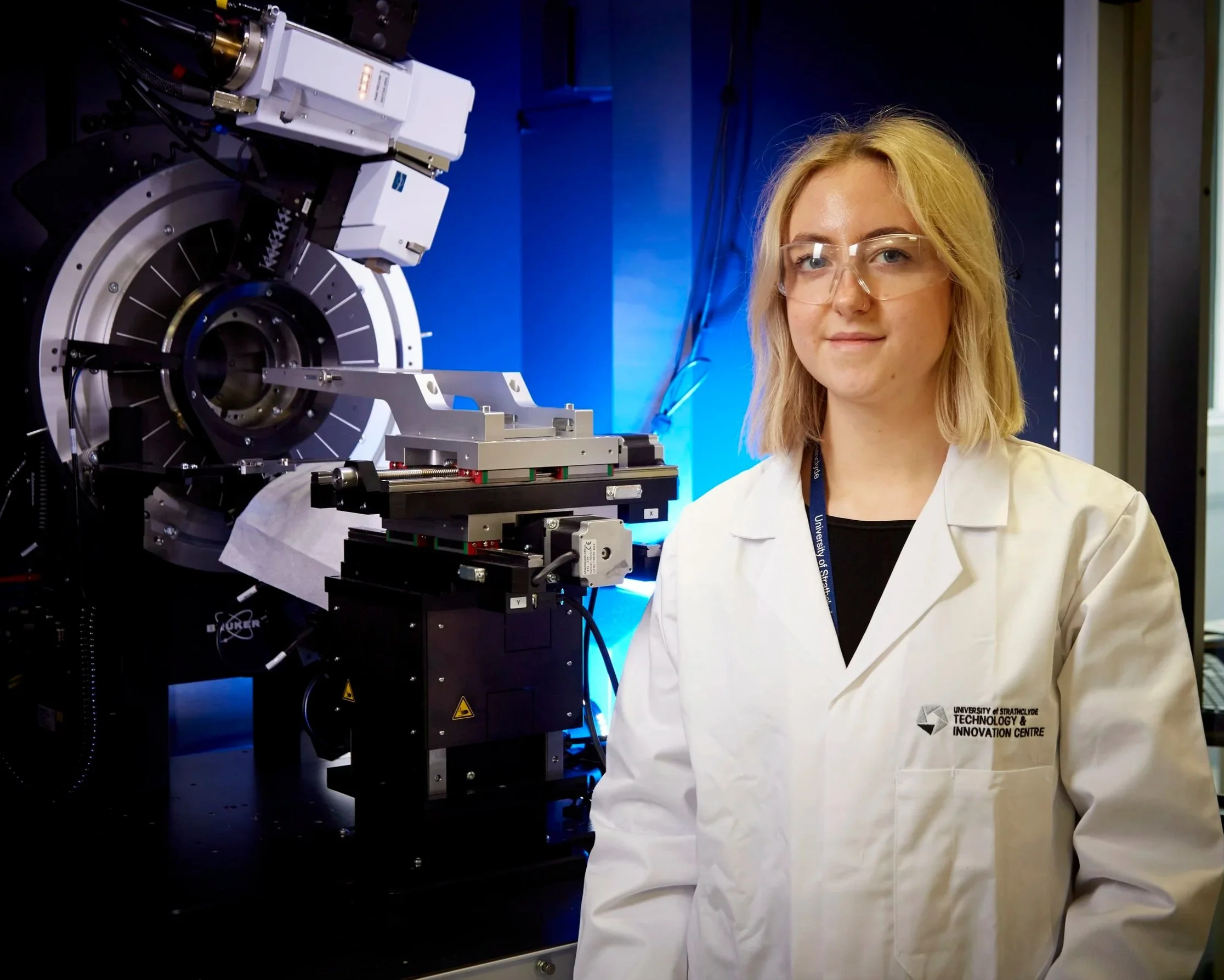 A young woman with blonde hair and safety glasses in a laboratory, standing next to scientific equipment, with a blue background and a white lab coat with a logo from the University of Strathclyde's Technology and Innovation Centre.