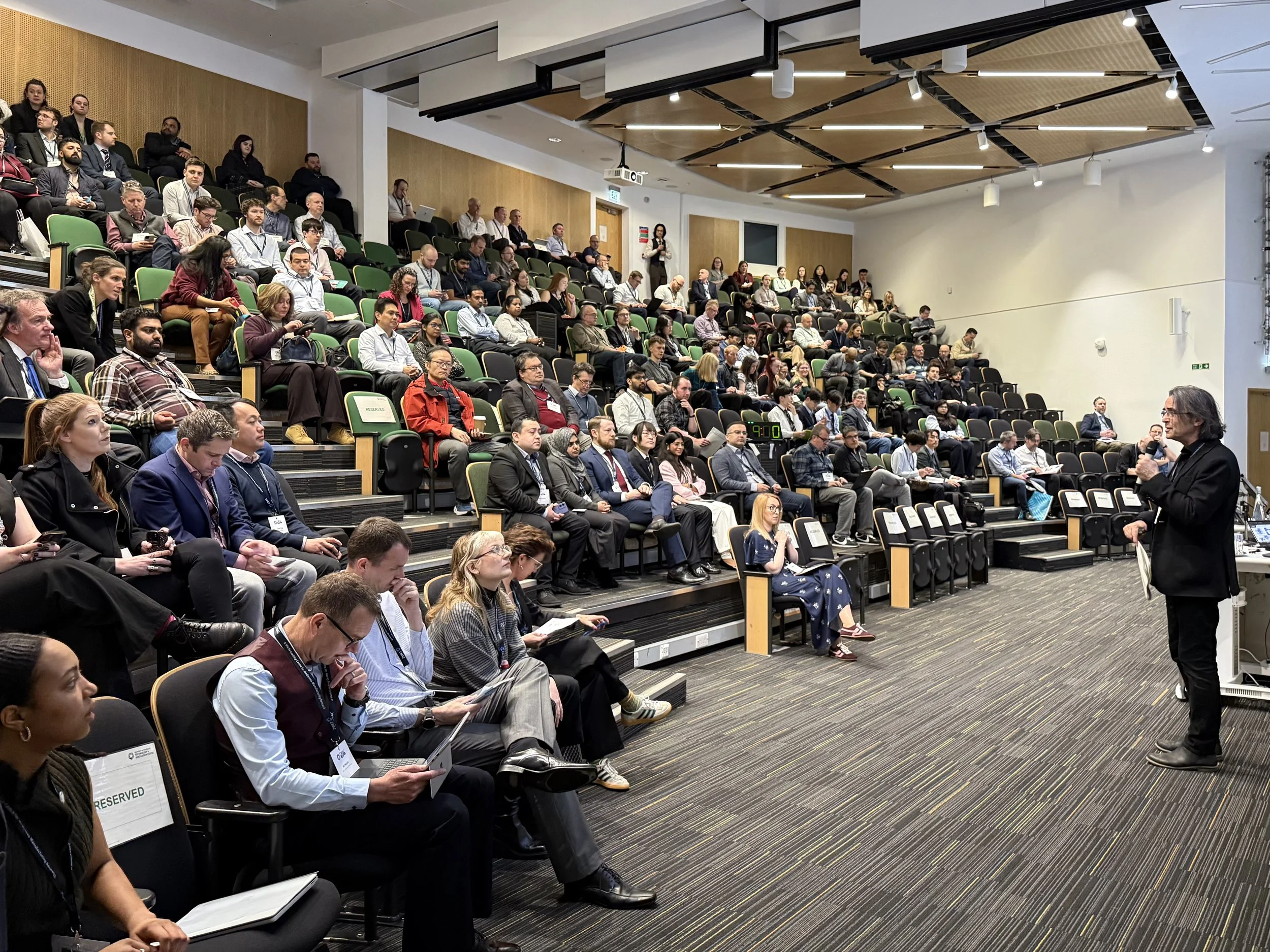 A large audience seated in tiered rows attending a conference or presentation. The room has a modern design with a wooden ceiling and a projector hanging from above. A speaker with glasses and long hair is speaking in front of the audience.