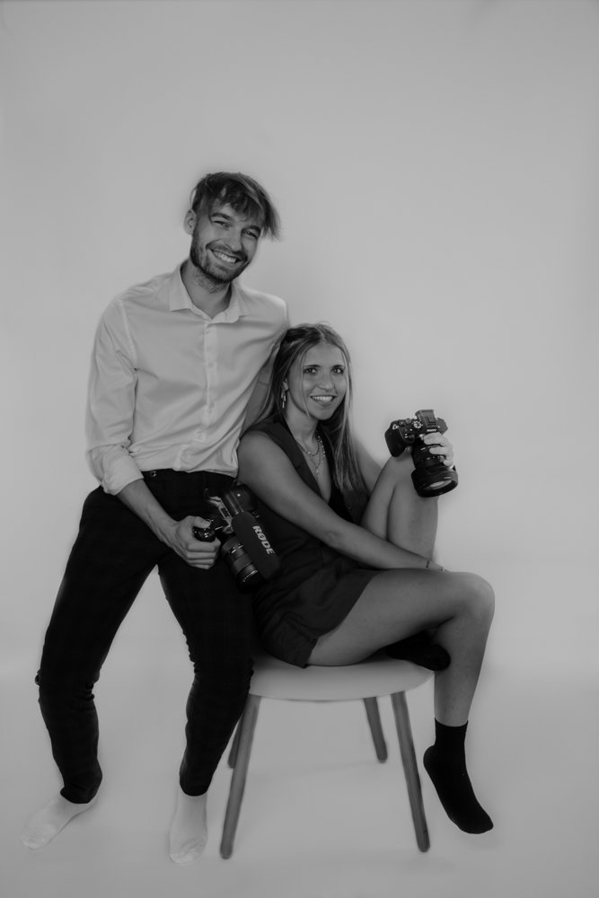 Duo de photographes posant avec humour en studio blanc, tenant chacun un appareil photo – l’équipe couple de LG photo & film.