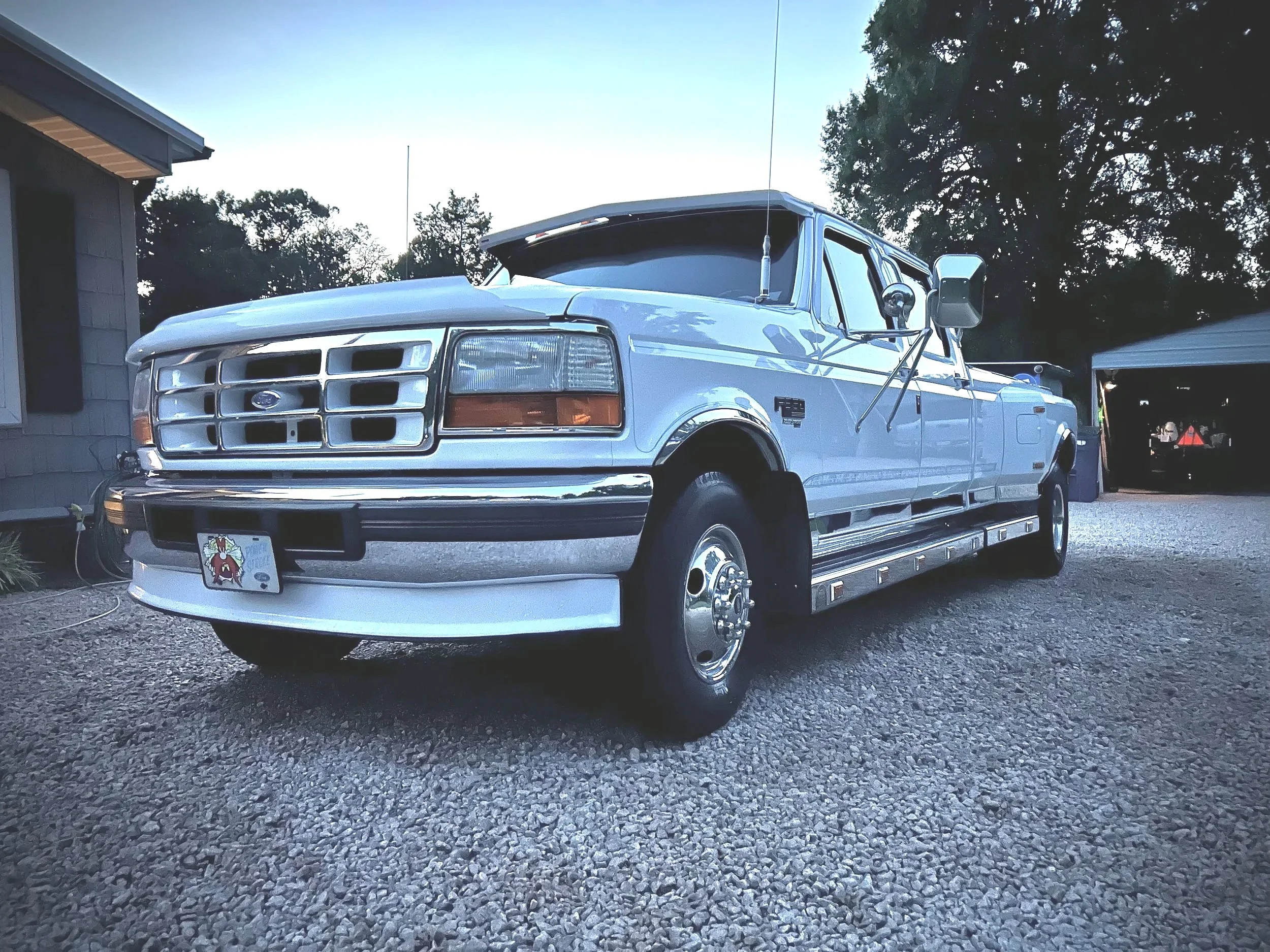 A vintage white Ford truck parked on a gravel driveway with a garage and trees in the background.