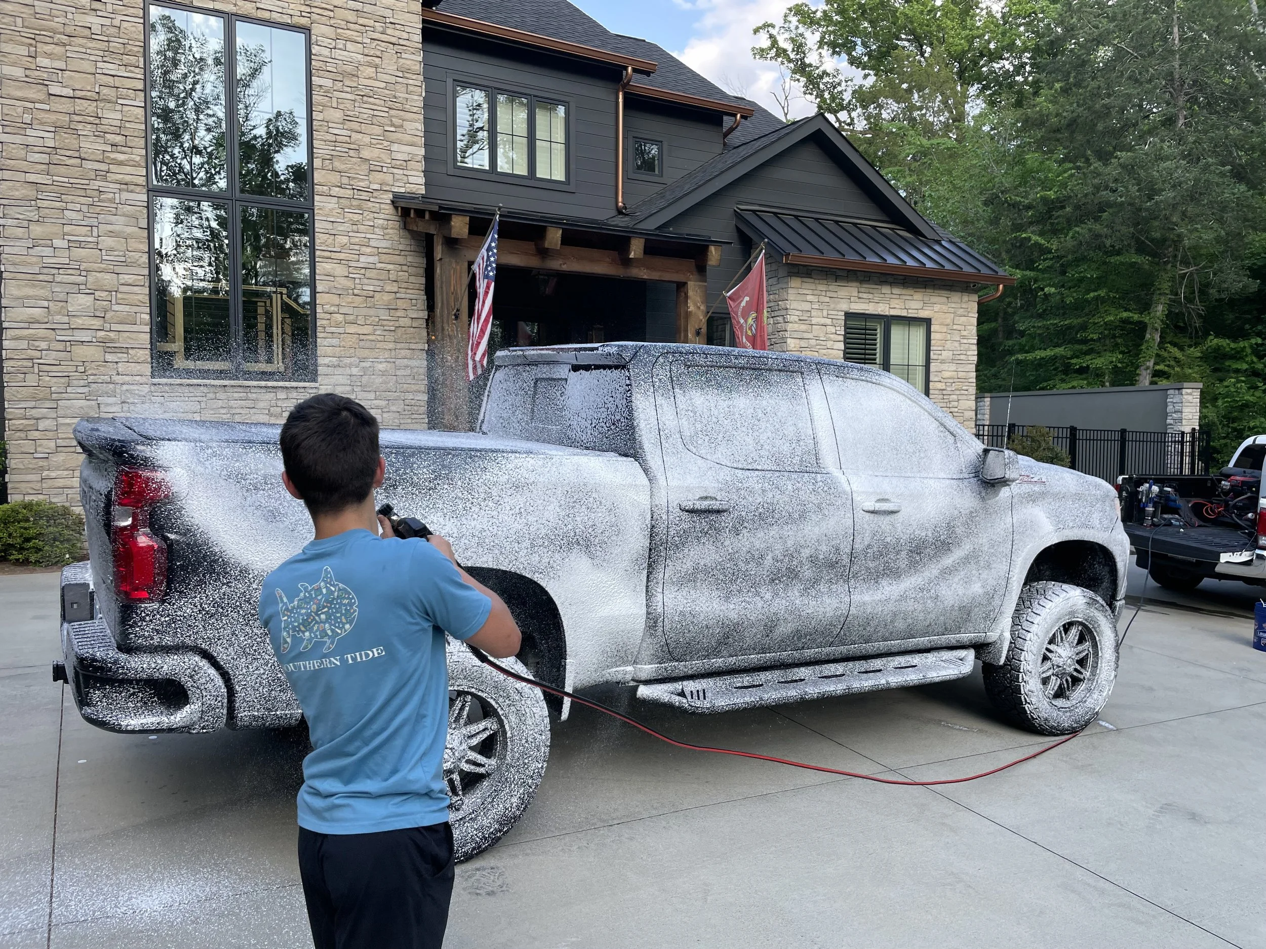 A young boy is pressure washing a large gray truck covered in soap suds in the driveway of a modern house with brick and gray siding. The house has American and military flags hanging outside.