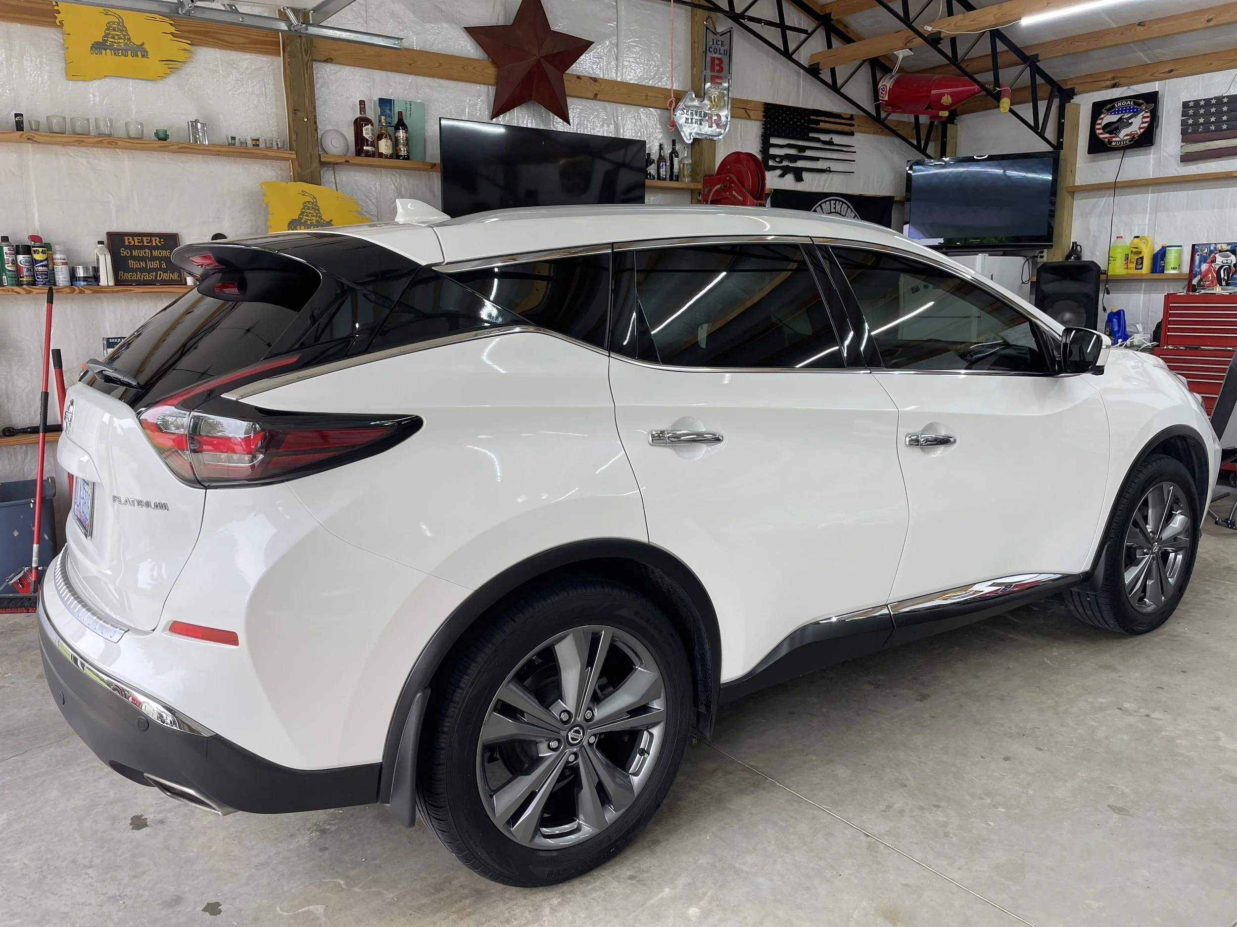 A white electric vehicle with a black roof and tinted windows parked inside a garage. The garage has shelves with bottles and tools, and various decorations including a red star and patriotic flags.