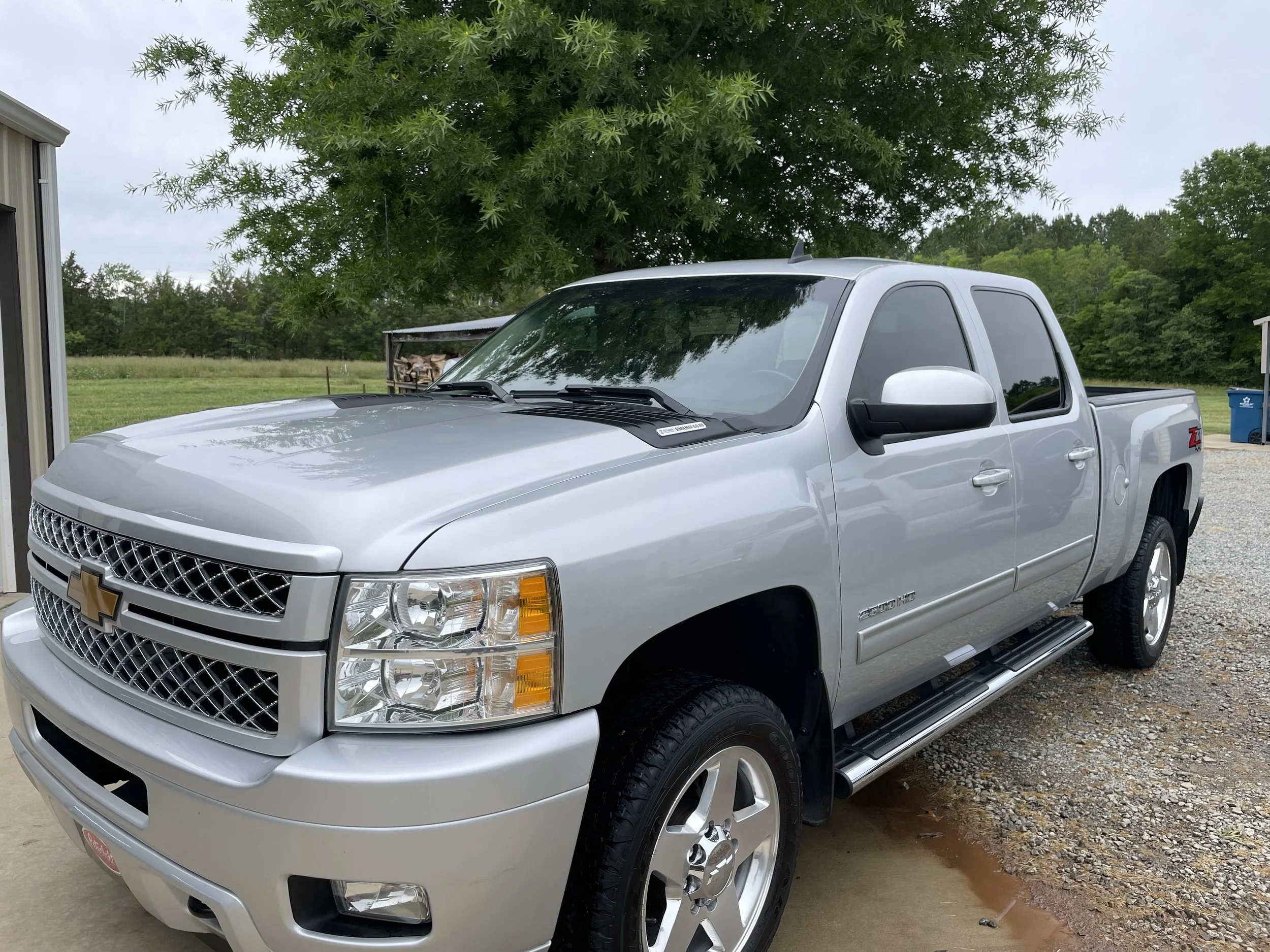 Silver Chevrolet Silverado pickup truck parked outdoors on a gravel surface, with a large green tree in the background.