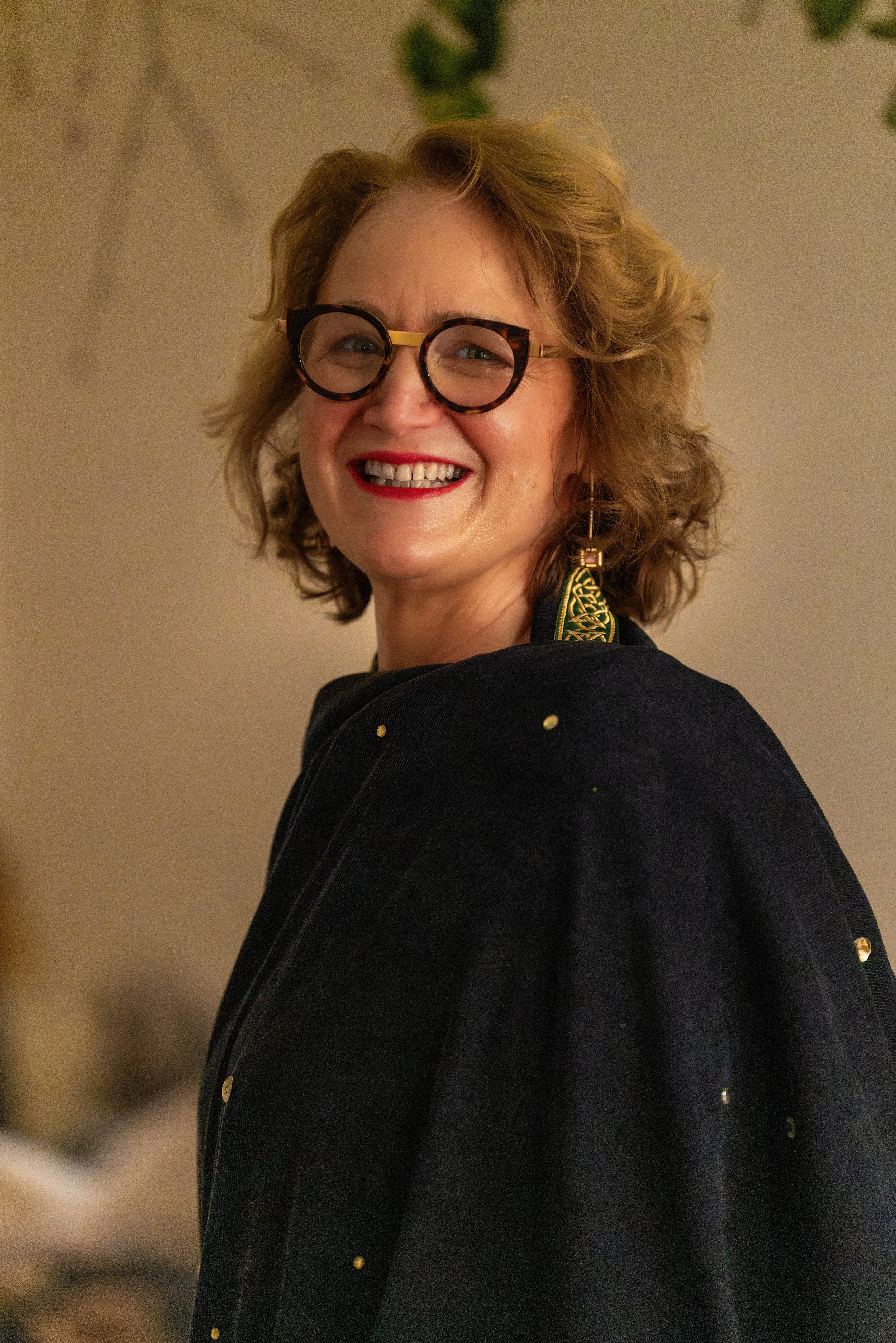 Smiling woman with curly hair, wearing glasses, red lipstick, a black jacket with gold embellishments, and gold earrings.