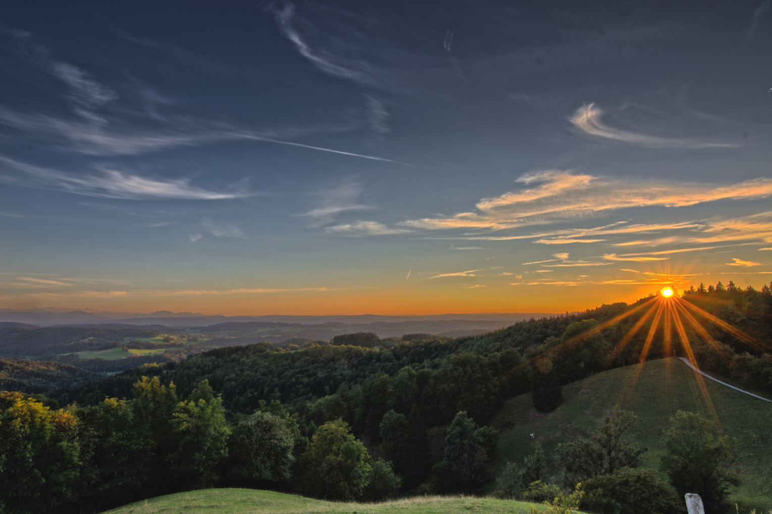 Sunset over rolling green hills and forested landscape with a partly cloudy sky.