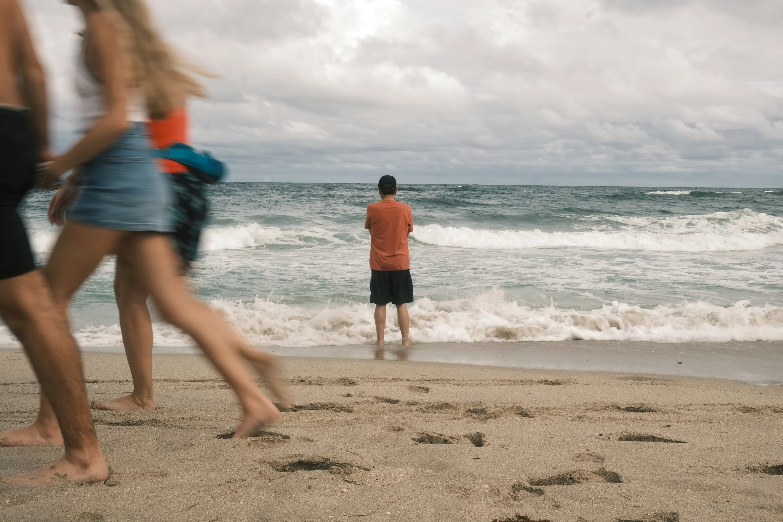 A person stands at the shoreline facing the ocean with feet in the water, while two other people walk along the beach in the foreground.