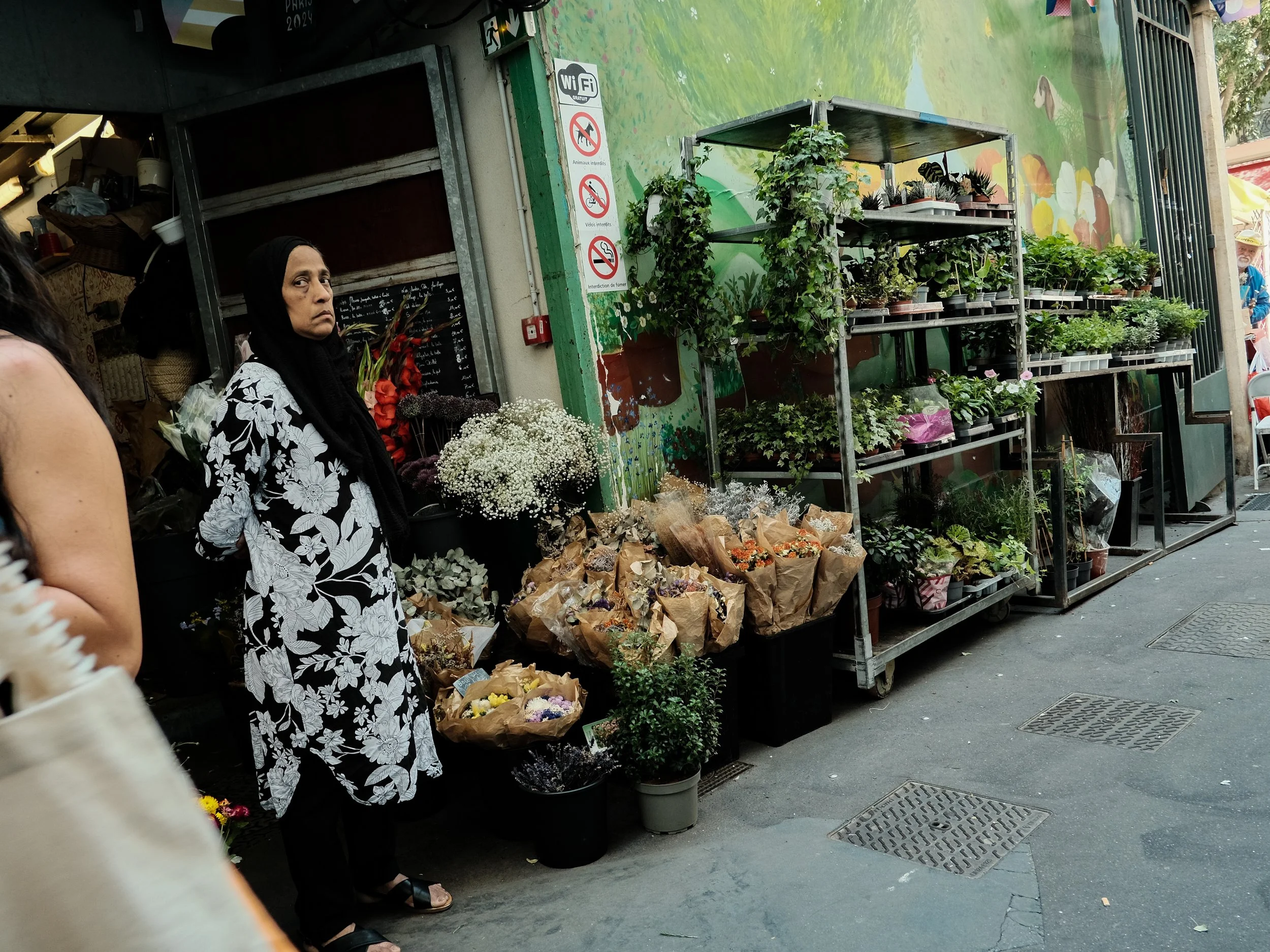 A woman standing outside a flower shop on a city sidewalk, surrounded by various potted plants and bouquets, with a mural painted on the wall behind her.