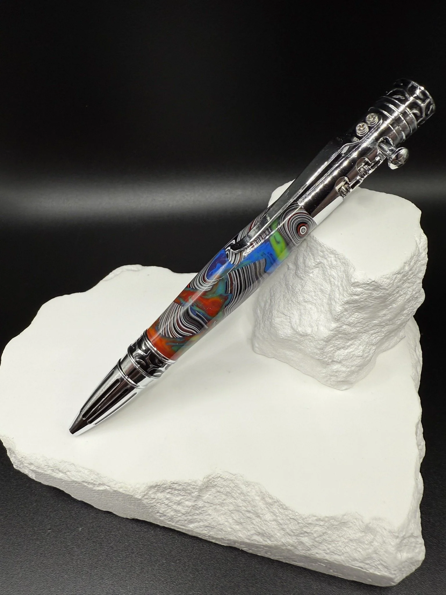 A colorful, patterned Mopar Fordite gear click pen resting on a white decorative rock and stand against a black background.
