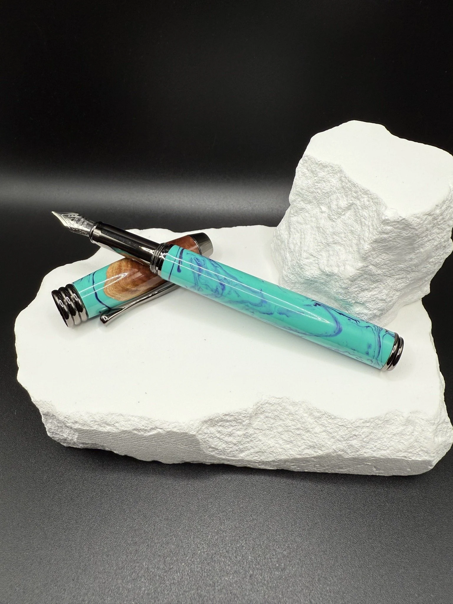 A turquoise marbled Red Mallee hybrid fountain pen with a black nib resting on a white stone slab, next to a rough white rock.