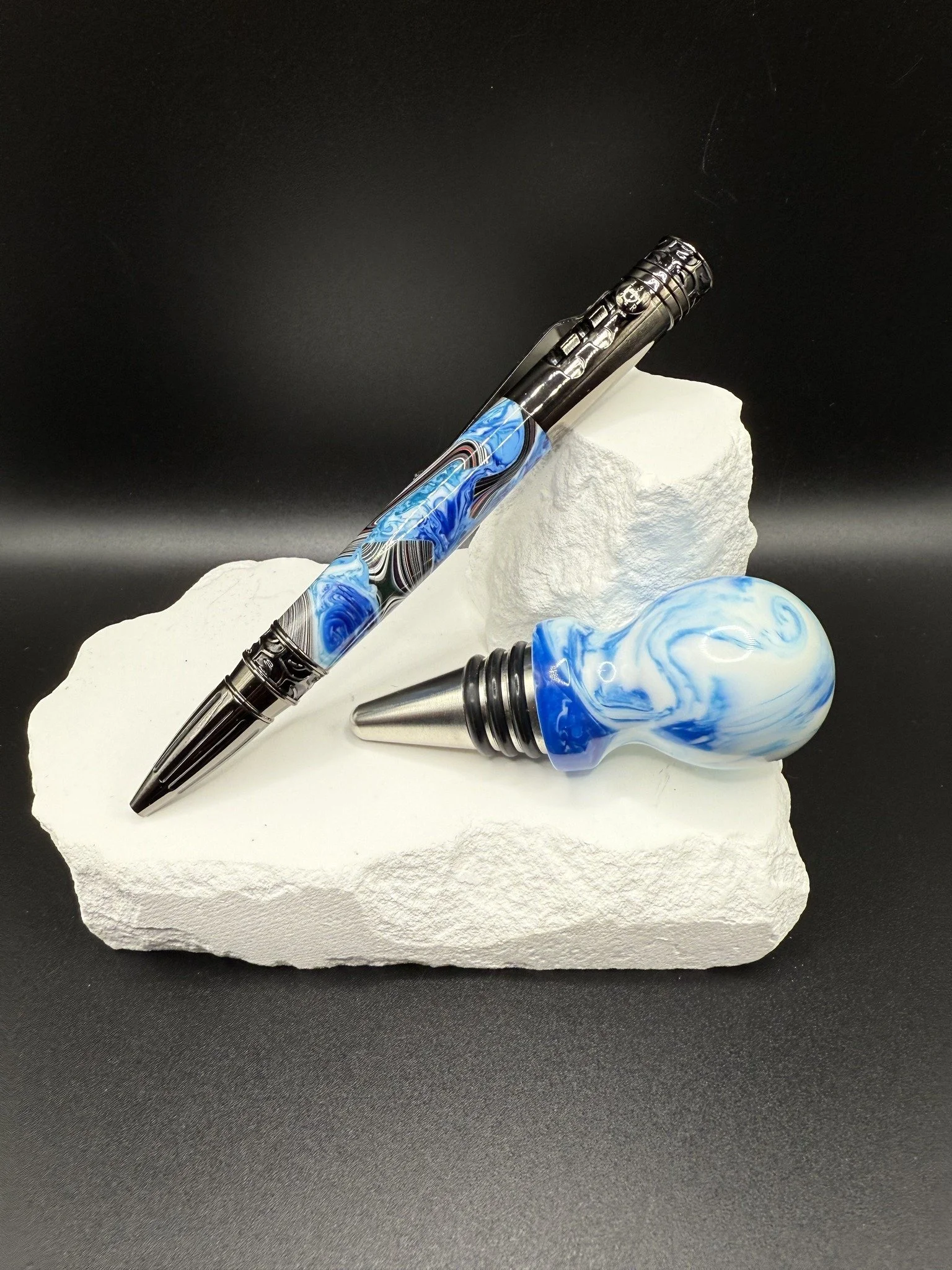 A pen and a bottle stopper with blue and white swirled marble designs resting on white textured rocks against a black background.