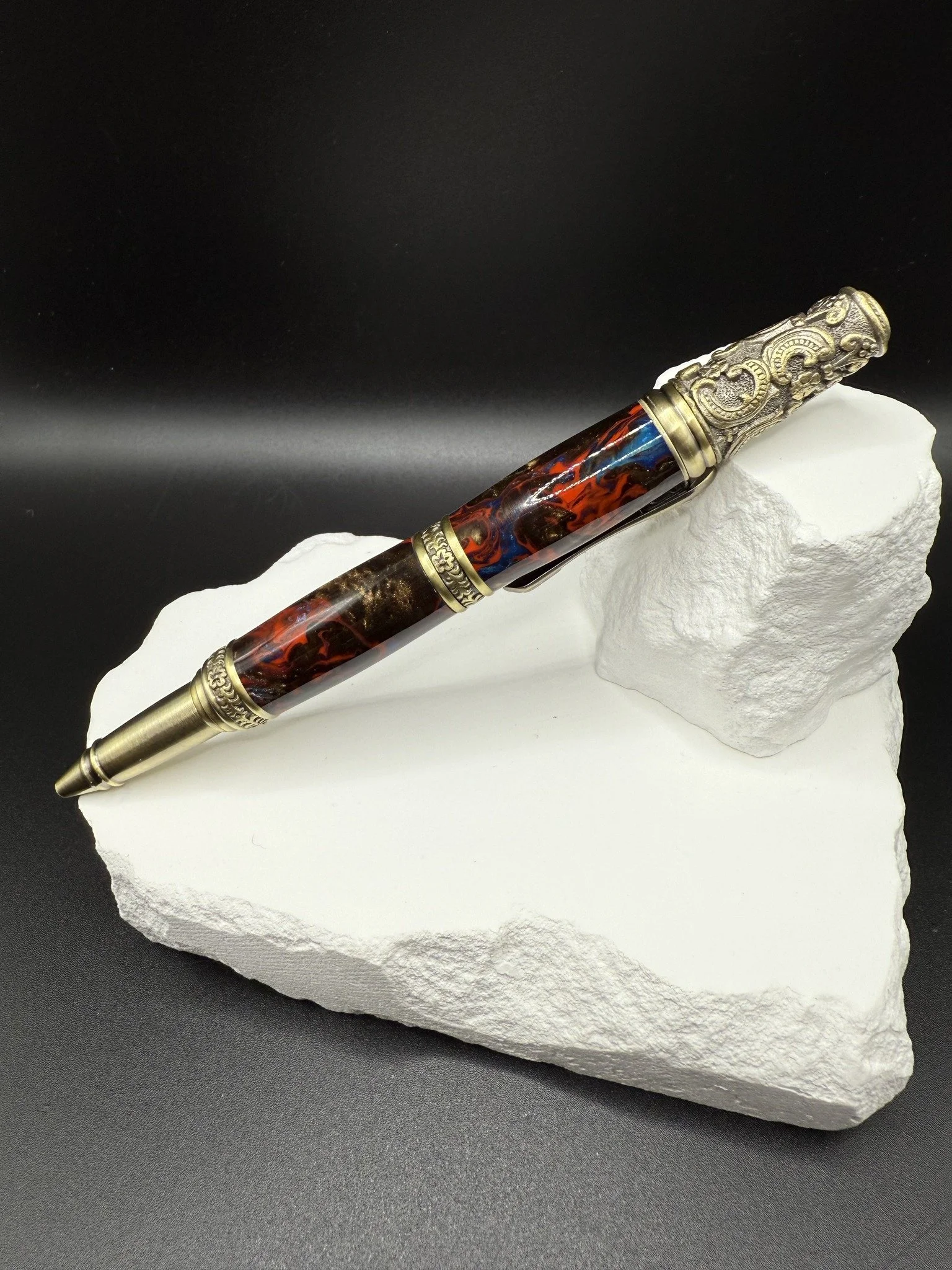 Acrylic decorative pen with ornate metallic and resin details resting on a white textured stone block against a black background.