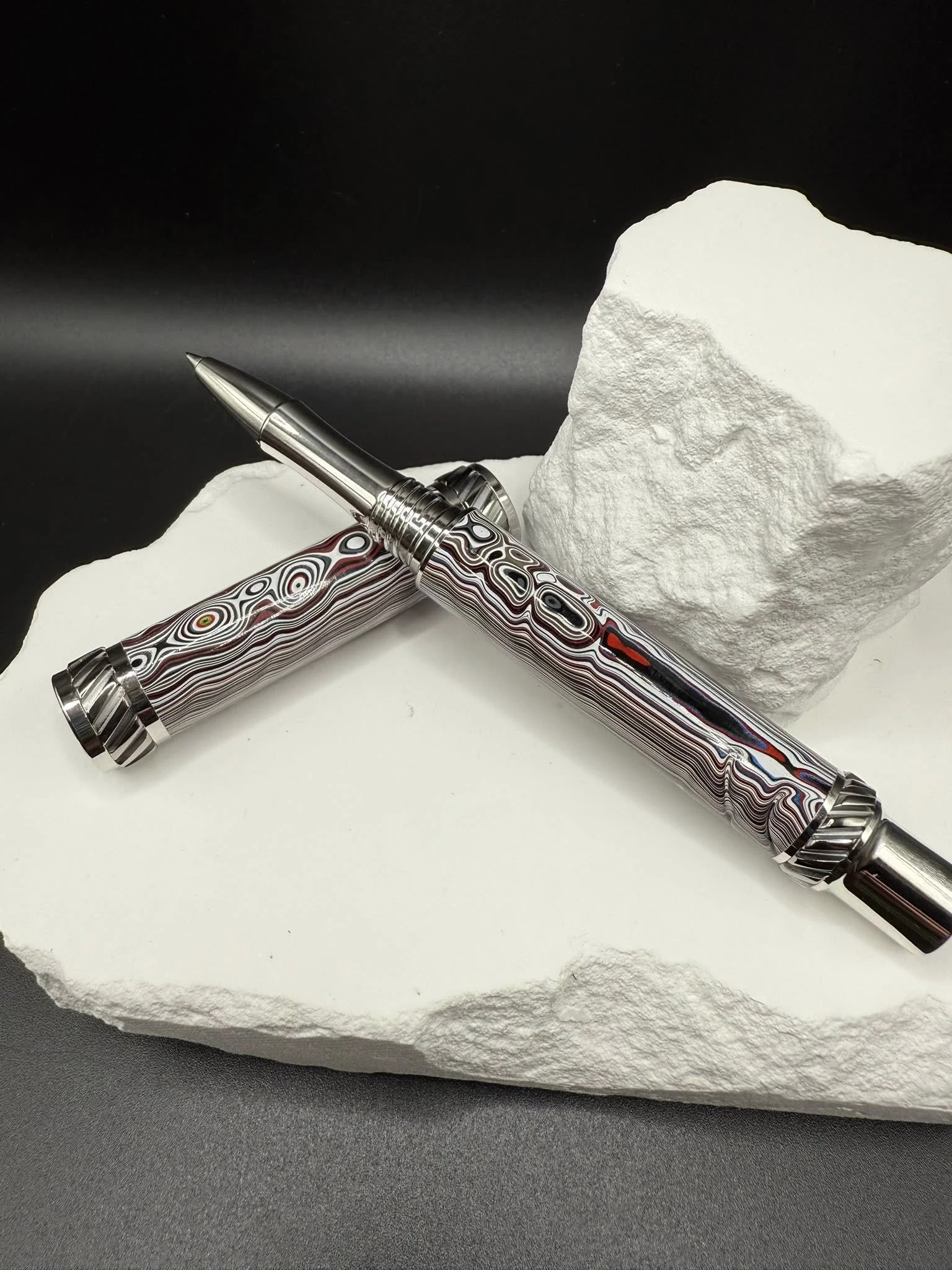 A shiny, patterned pen with a swirling multicolored design is resting on a rough white stone, with another similar pen lying beside it, on a black and white surface.