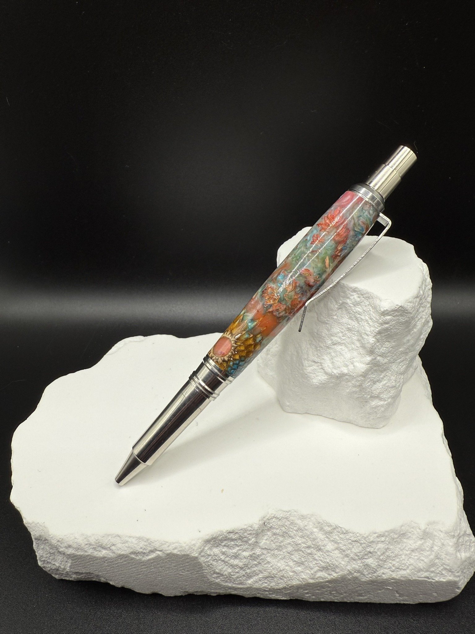 A multicolored, Wildflower and acrylic pen resting on a white textured block of material against a black background.