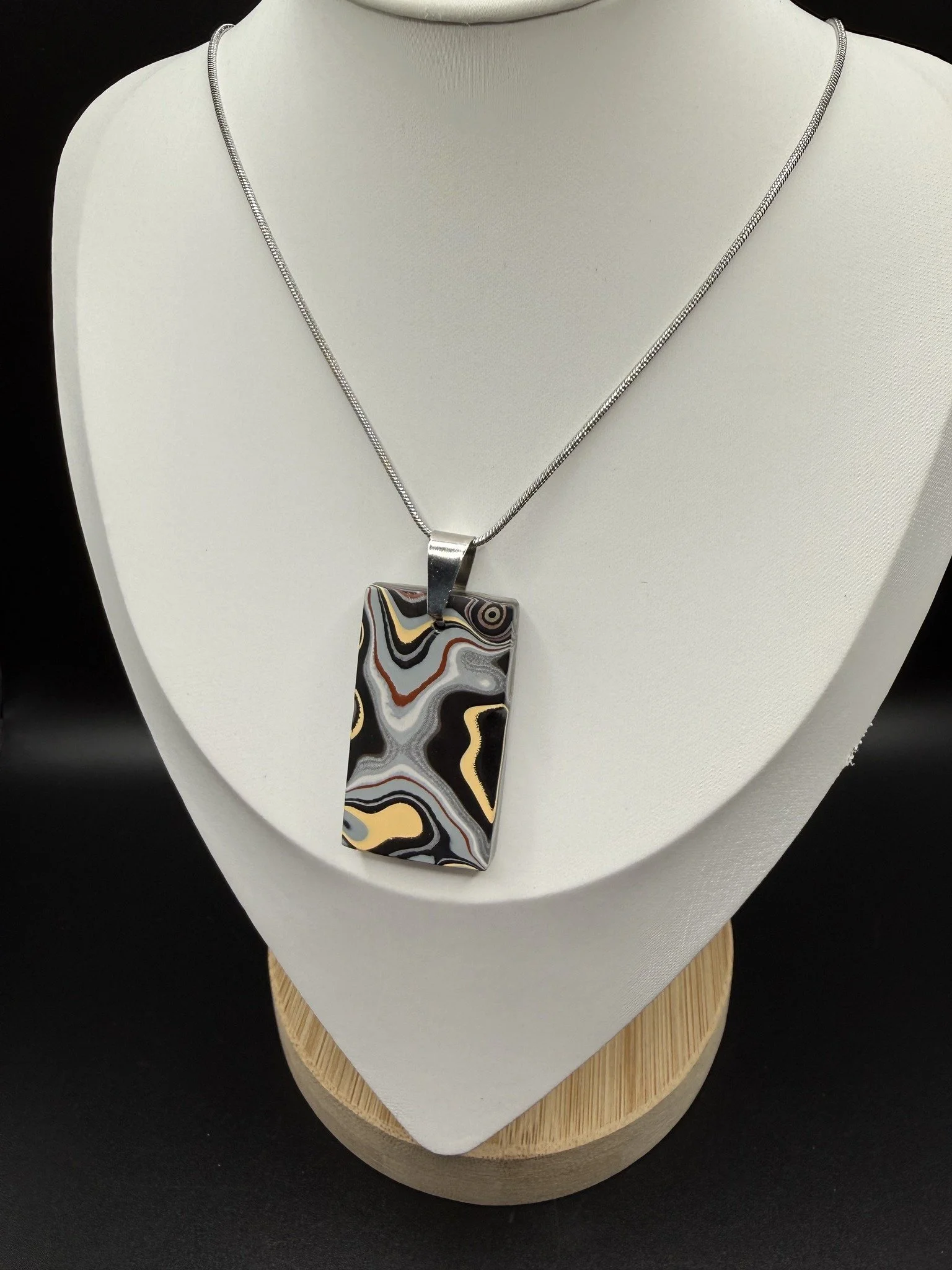 Fordite Necklace with a rectangular pendant featuring black, white, yellow, and gray swirled patterns, displayed on a white bust stand.