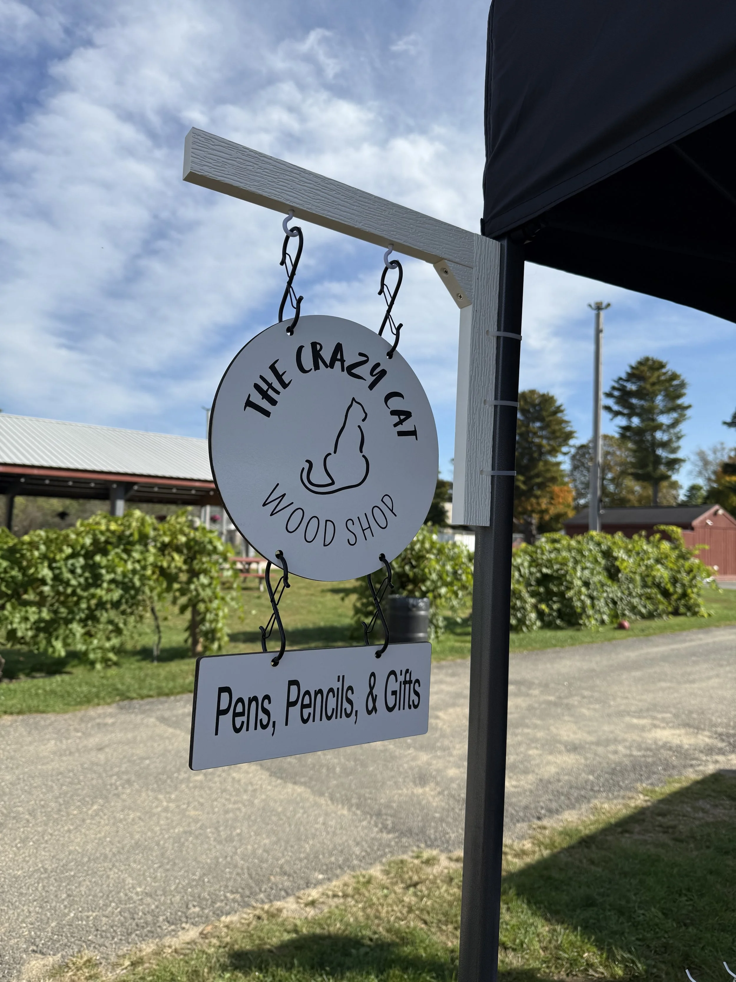 Sign for The Crazy Cat Wood Shop, offering pens, pencils, and gifts, hanging on a display stand outdoors with trees and a blue sky in the background.