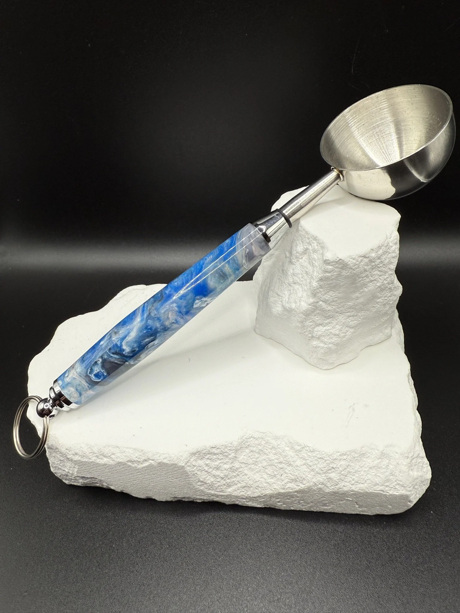 A metallic acrylic coffee scoop resting on a white textured stone with a blue swirled handle, against a black background.