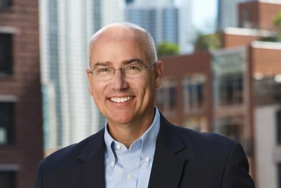 Headshot of a bald man with glasses, smiling, wearing a light blue dress shirt and dark blazer, with a cityscape in the background.