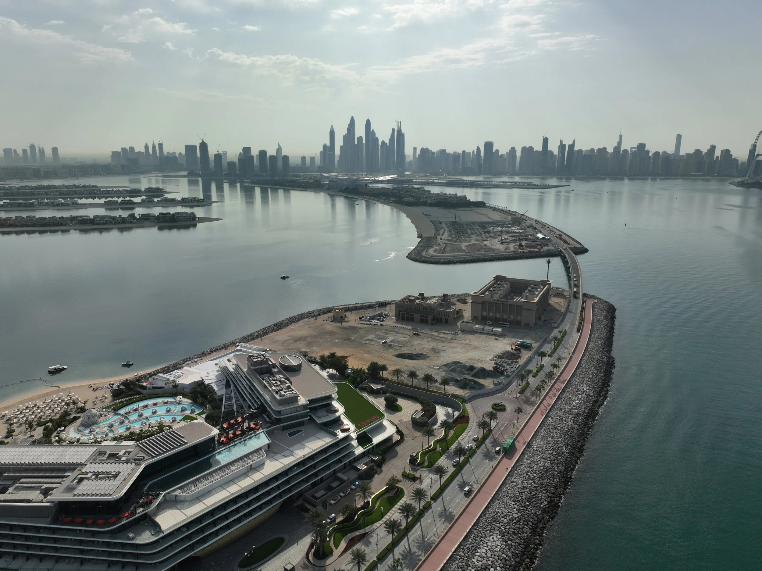 Aerial view of a city skyline with high-rise buildings, water, and a shoreline with a curved road, a large building, a hotel with a pool, and distant construction sites in the background.