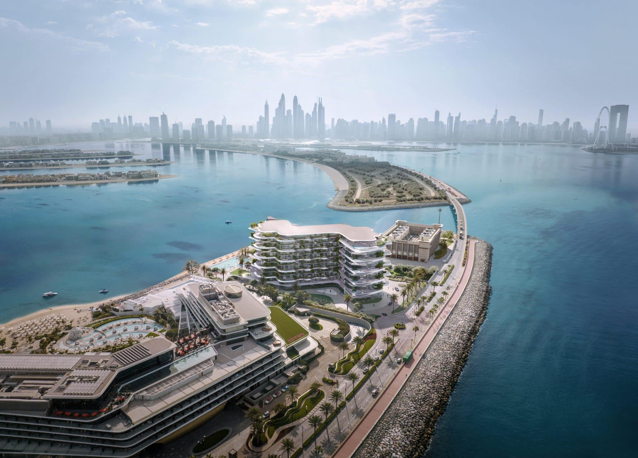 Aerial view of a modern waterfront development with a curved building, palm trees, and a sea wall, with the Dubai skyline featuring tall skyscrapers and the Ferris wheel in the distance.