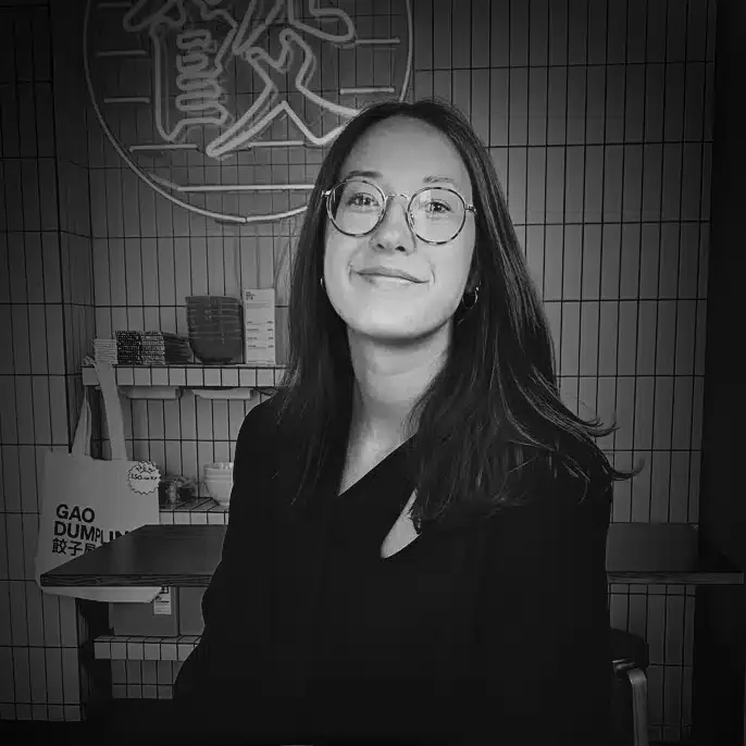 A woman with long dark hair, glasses, and a black top, smiling in a restaurant or cafe with a neon sign and shelving behind her.