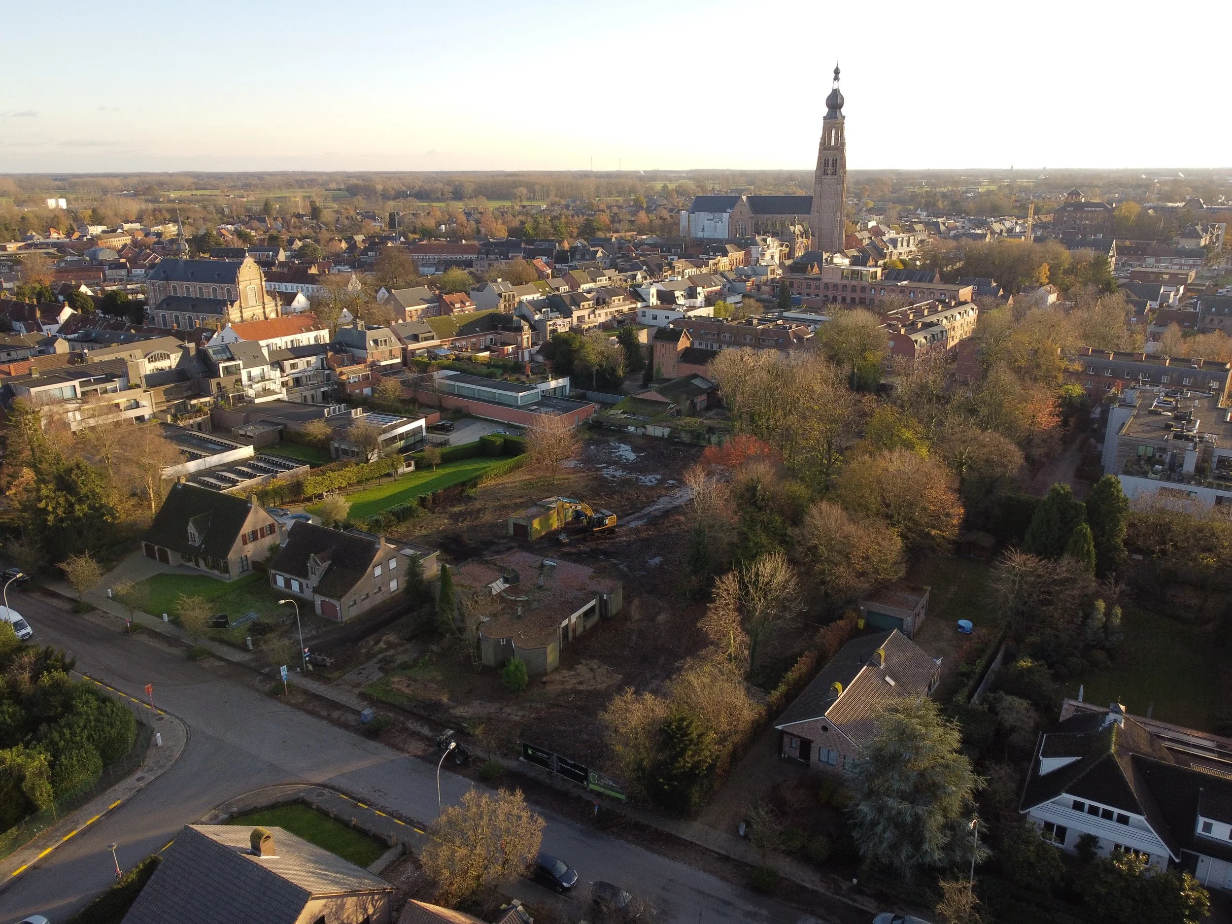 Aerial view of a city with a mix of residential and historic buildings, including a prominent tall church tower, and a construction site in the foreground with trees and houses.