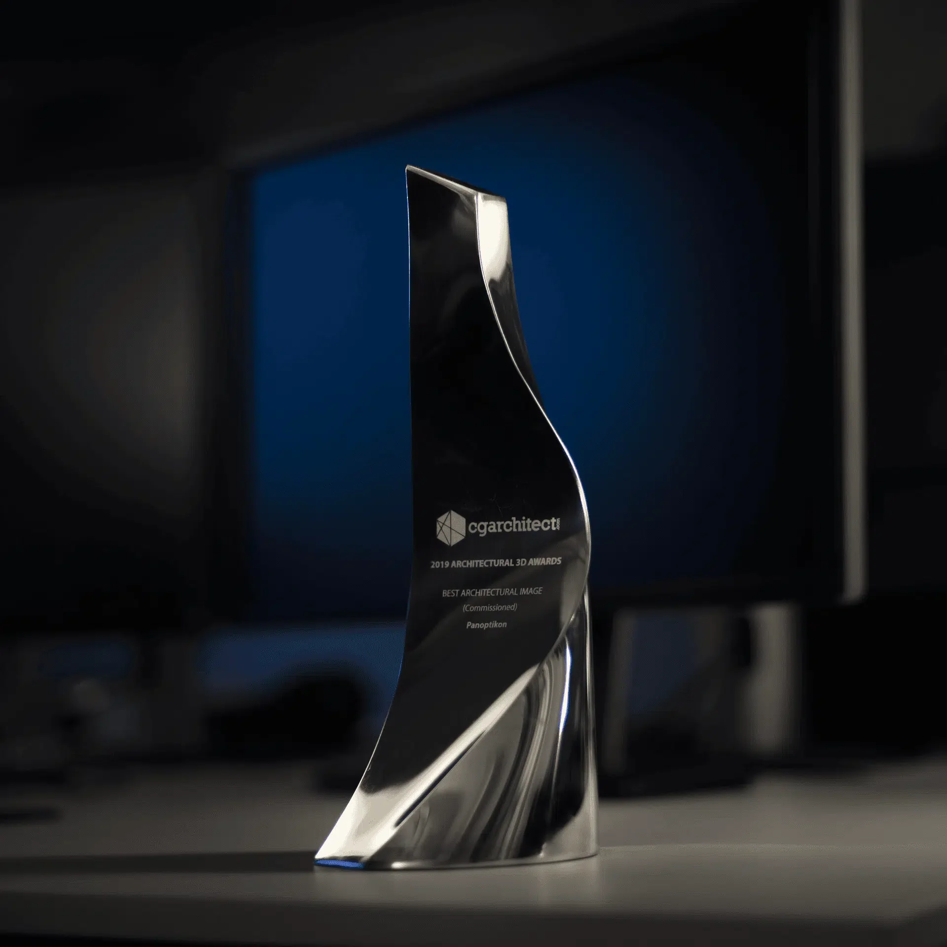 Shiny, abstract architectural award trophy with engraved text, placed on a desk with a computer monitor in the background.